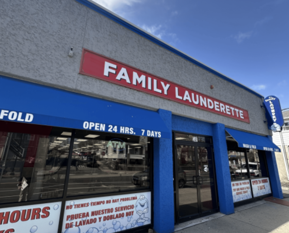 Exterior of a family laundromat with blue and gray walls, a red sign with white lettering that says "FAMILY LAUNDERETTE," and blue awnings advertising 24-hour service and no wait times.