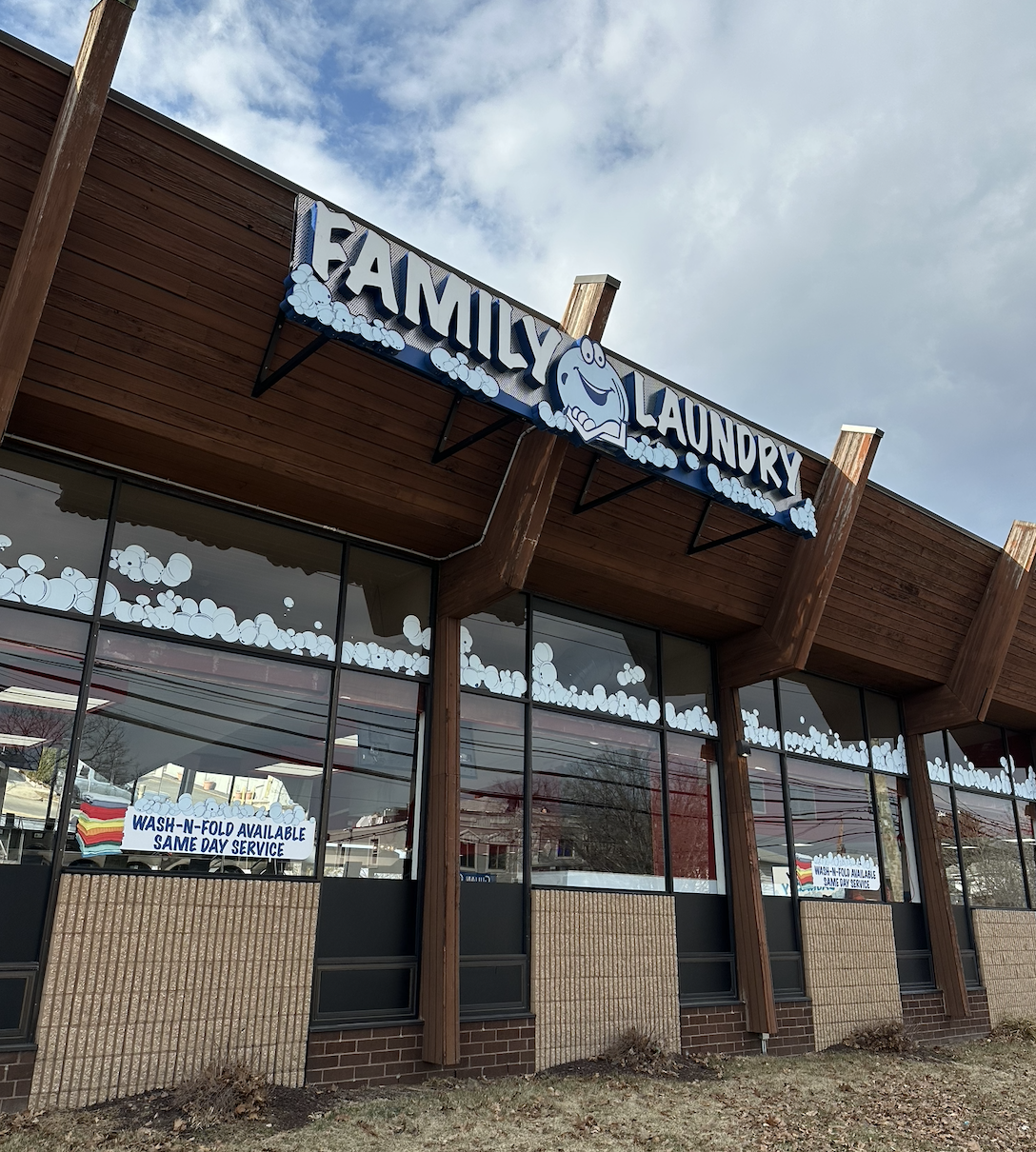 Exterior of a family laundry business with a sign that reads 'Family Laundry' featuring a cartoon smiling washing machine. The windows have soap bubble decorations, and there are signs indicating wash-and-fold service is available on the same day.