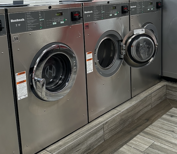 Three stainless steel commercial laundry machines with front-loading doors in a row, located on a wooden floor.