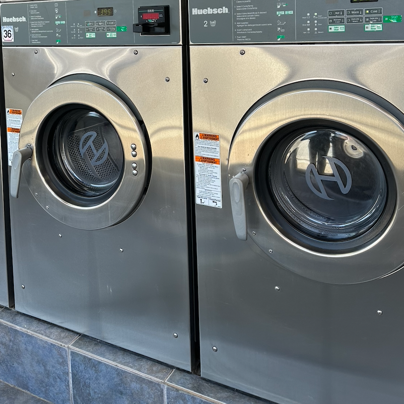 Two front-loading commercial washing machines made of stainless steel, placed side by side in a laundry area.