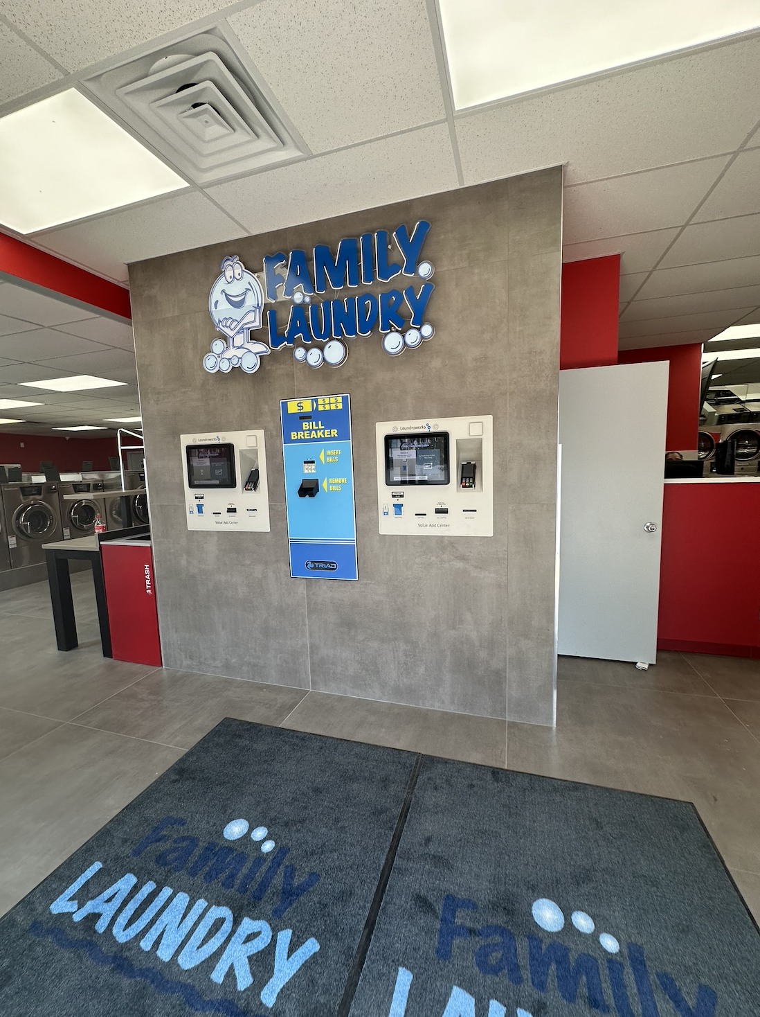 A laundromat with a sign for 'Family Laundry' and two coin-operated washers. There is a bill breaker machine between the washers, and a laundry mat with a blue mat on the floor.