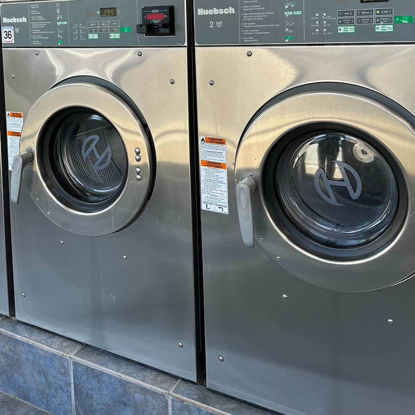 Two front-loading washing machines in a laundromat, made of stainless steel, with digital control panels and glass doors showing the washing drums inside.
