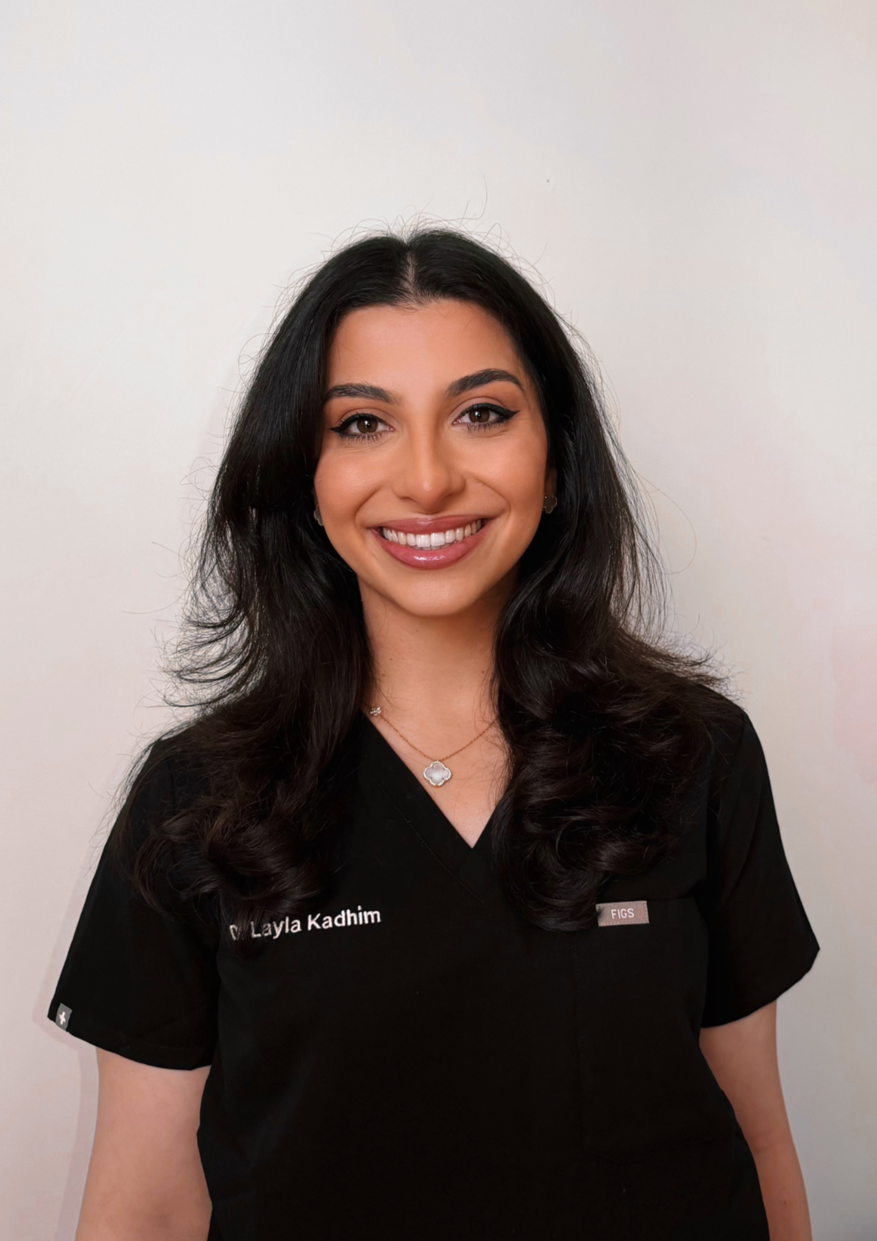 A woman with long dark hair smiling, wearing a black medical uniform with the name tag 'Dr. Layla Kadhim' and small earrings, standing against a plain white background.