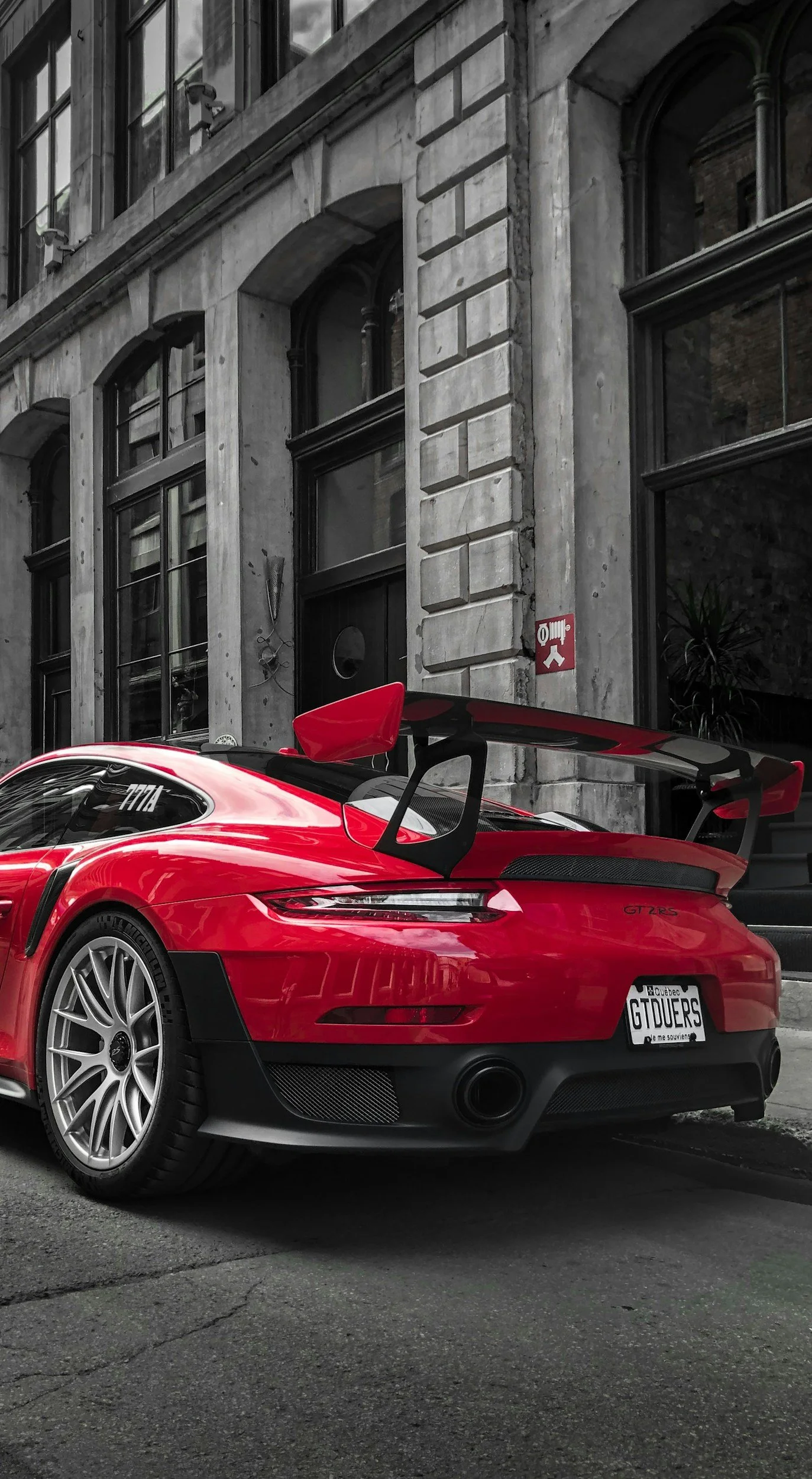 Red sports car parked in front of a gray brick building with large windows, with a Quebec license plate that reads 'GTDWERS'.