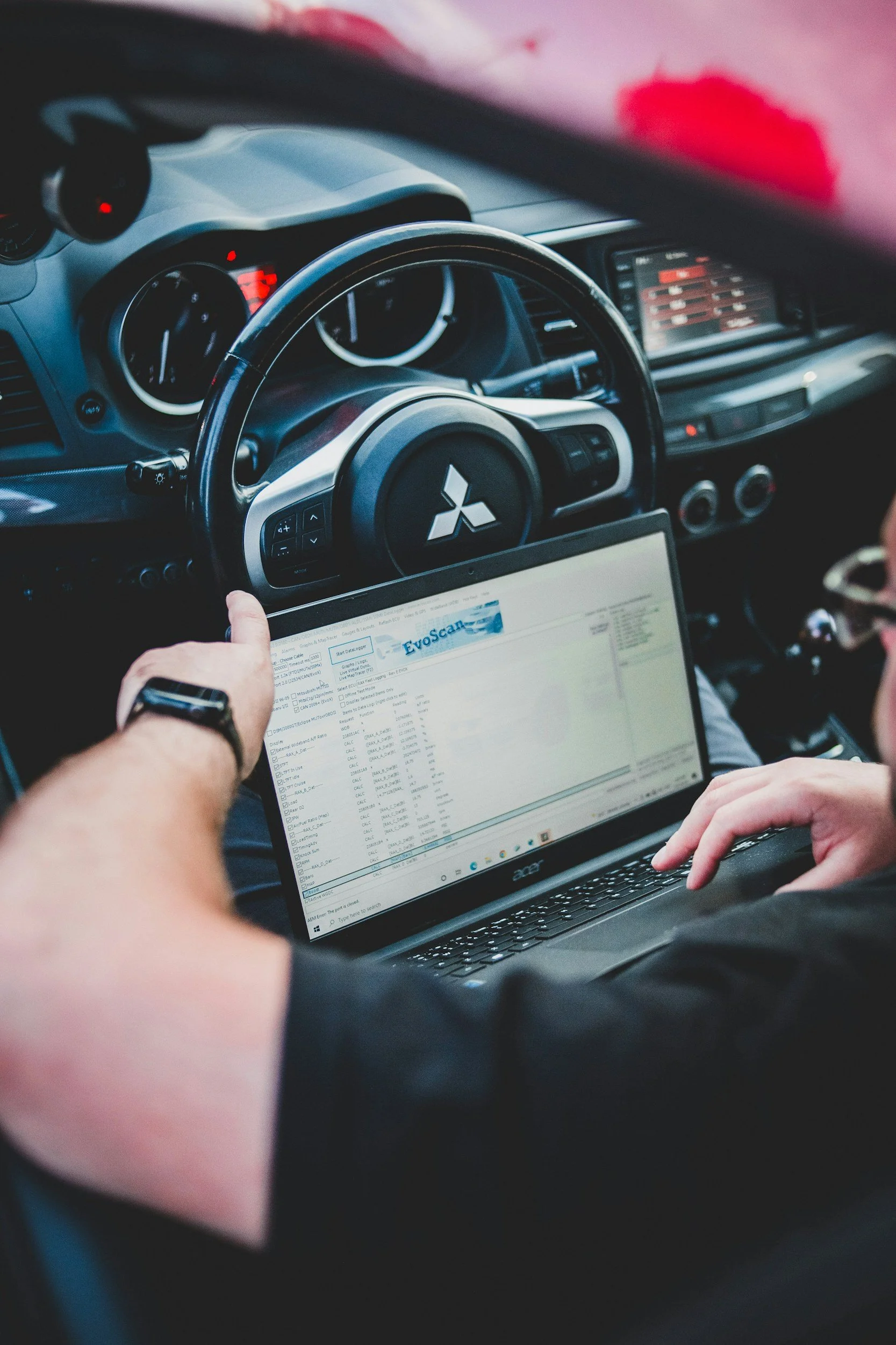 Inside a vehicle, a person is holding a laptop showing a software interface, with another person operating the vehicle's steering wheel, which has a Mitsubishi logo.