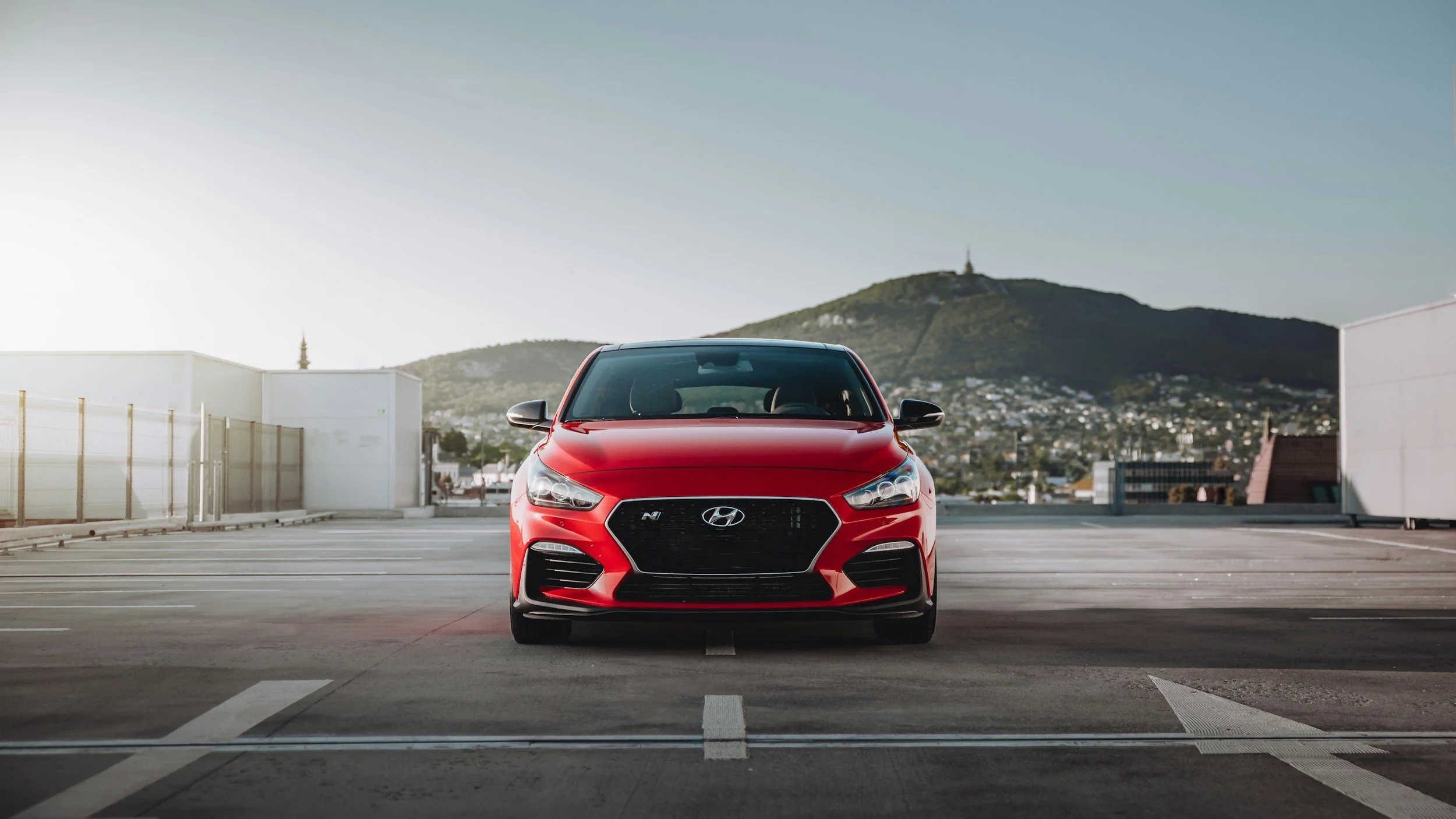 Red Hyundai Veloster N parked in an empty rooftop parking lot with city buildings and a mountain in the background.