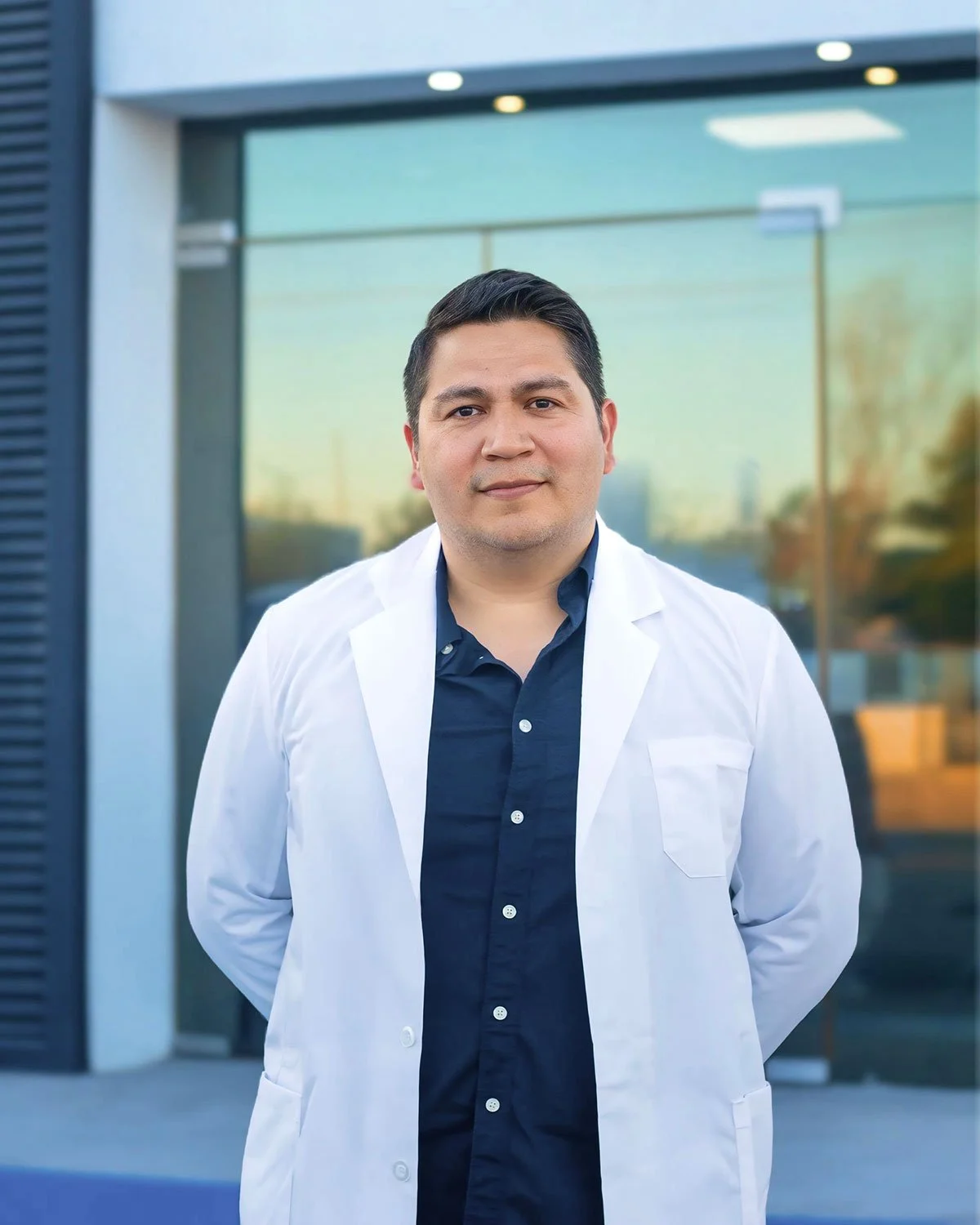 Un hombre con ropa formal y bata blanca frente a un edificio de vidrio con reflejo de árboles y cielo al atardecer.