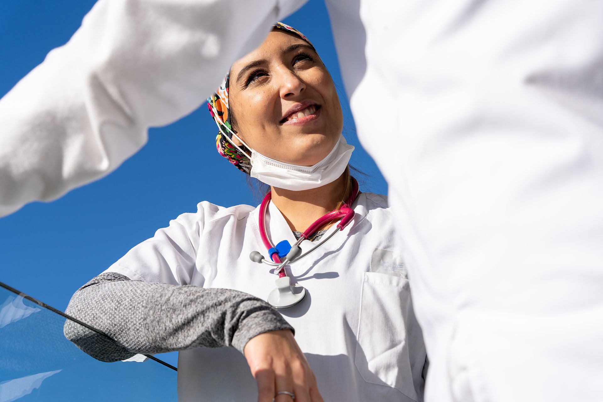 Una enfermera con uniforme blanco y estetoscopio, con una mascarilla bajada, mira hacia arriba y sonrie bajo el cielo azul.