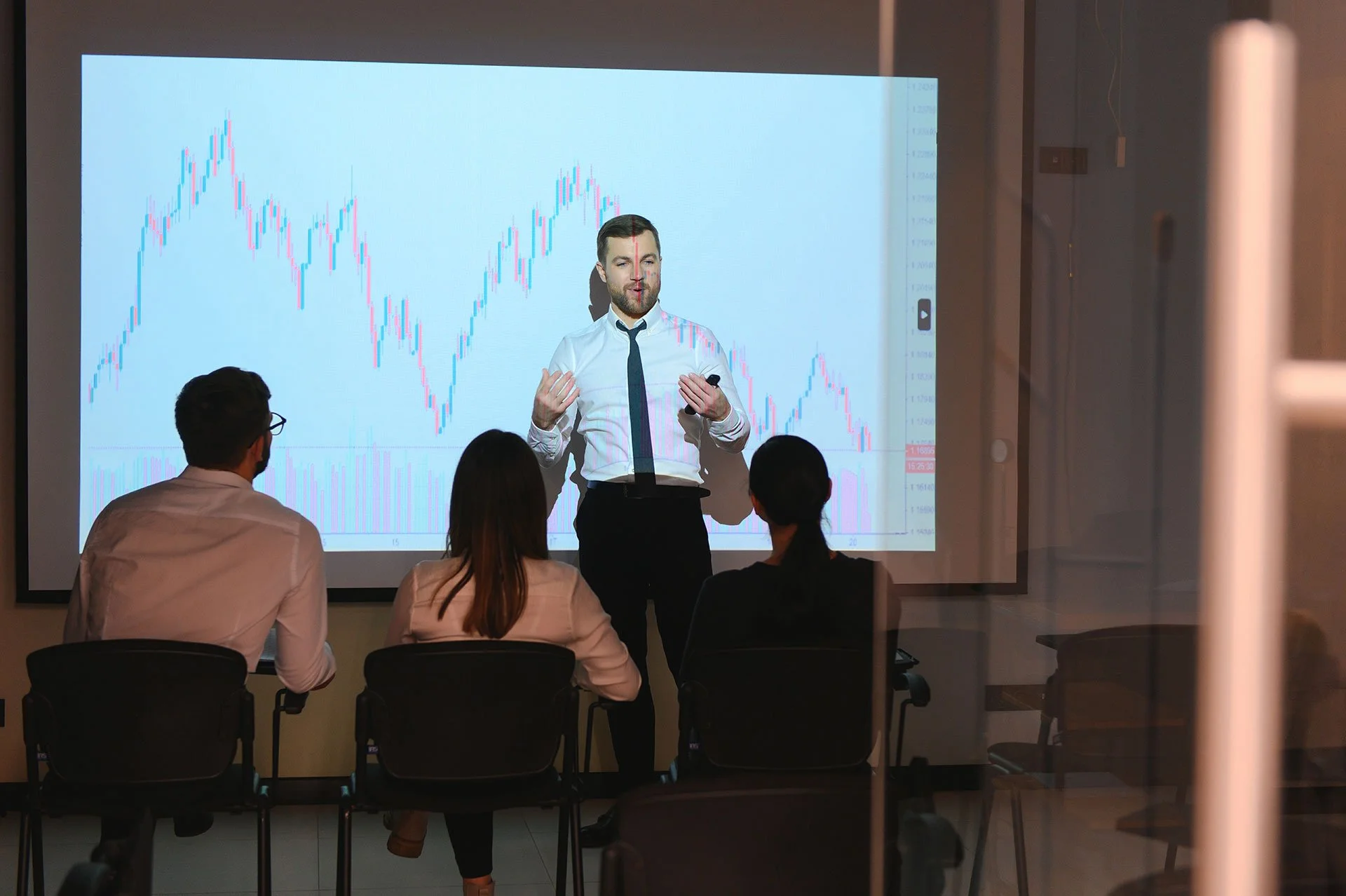 Hombre dando una presentación sobre gráficos de precios a un grupo de personas en una sala de conferencias.