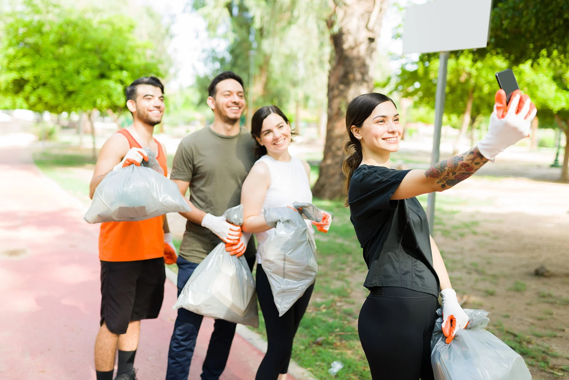 Grupo de cuatro personas participando en una actividad de limpieza en un parque, con árboles verdes y caminos pavimentados, sosteniendo bolsas de basura y usando guantes de limpieza.