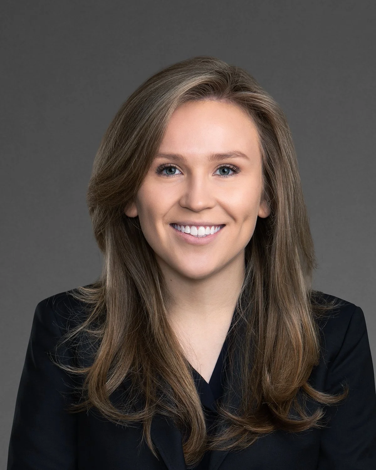 Headshot of a smiling woman with long wavy brown hair, blue eyes, wearing a black blazer, against a gray background.