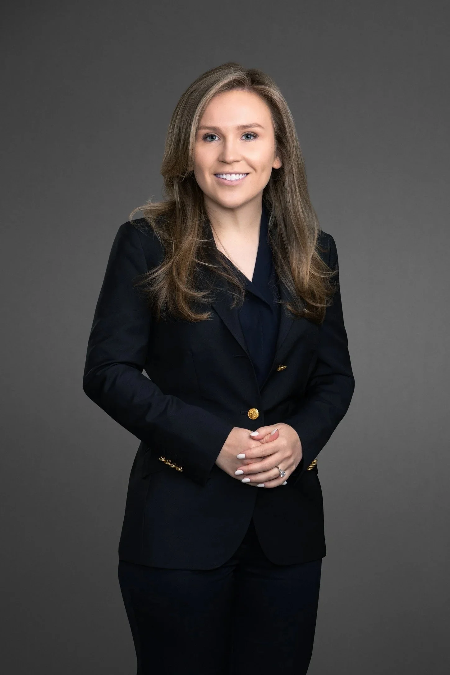 A professional woman with long, wavy brown hair, wearing a black blazer with gold buttons, standing against a plain gray background.
