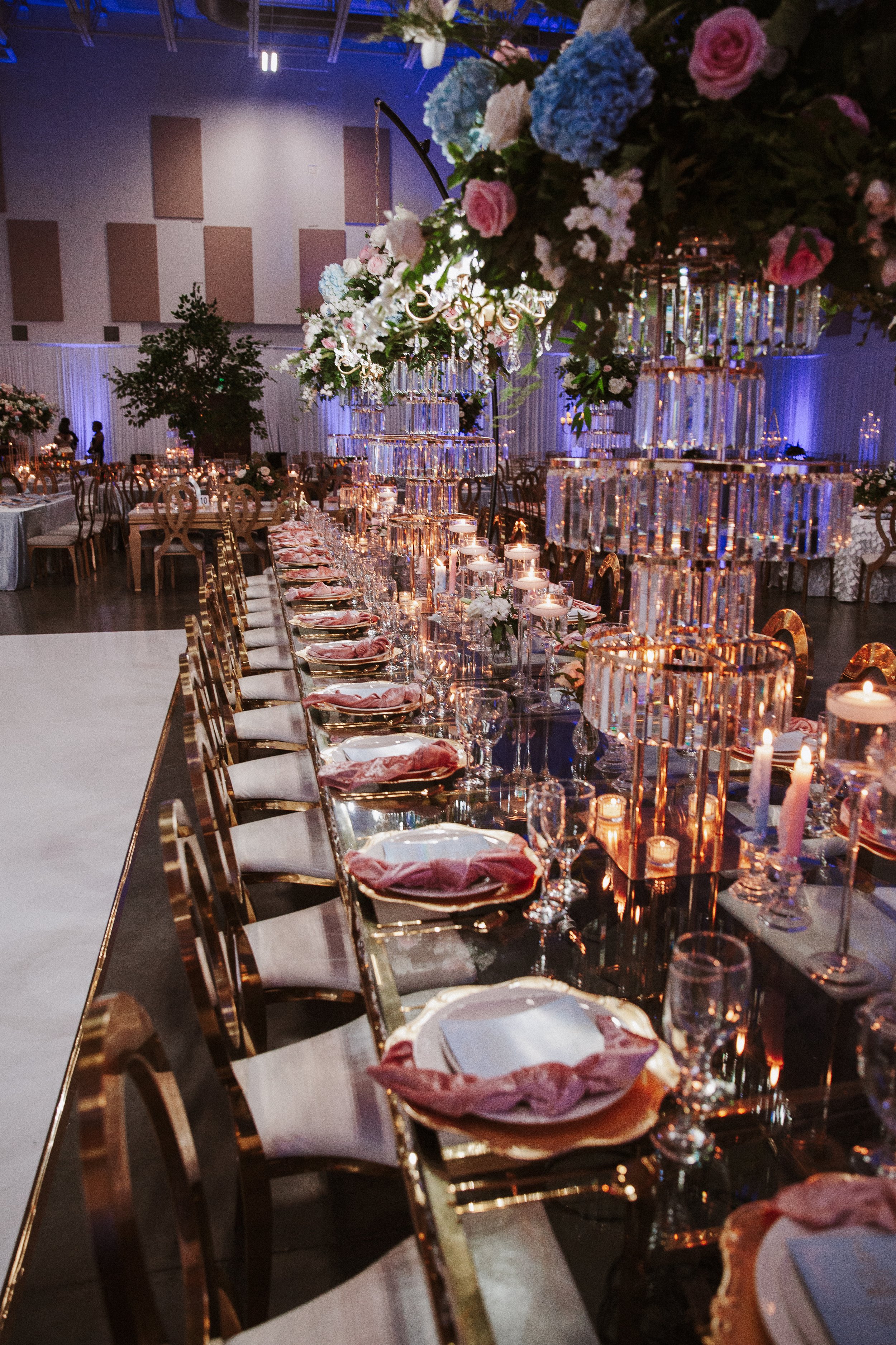 Elegant banquet hall decorated for a wedding reception or formal event, featuring a long table with gold chairs, floral centerpieces, and candles, with draped walls and hanging chandeliers in the background.