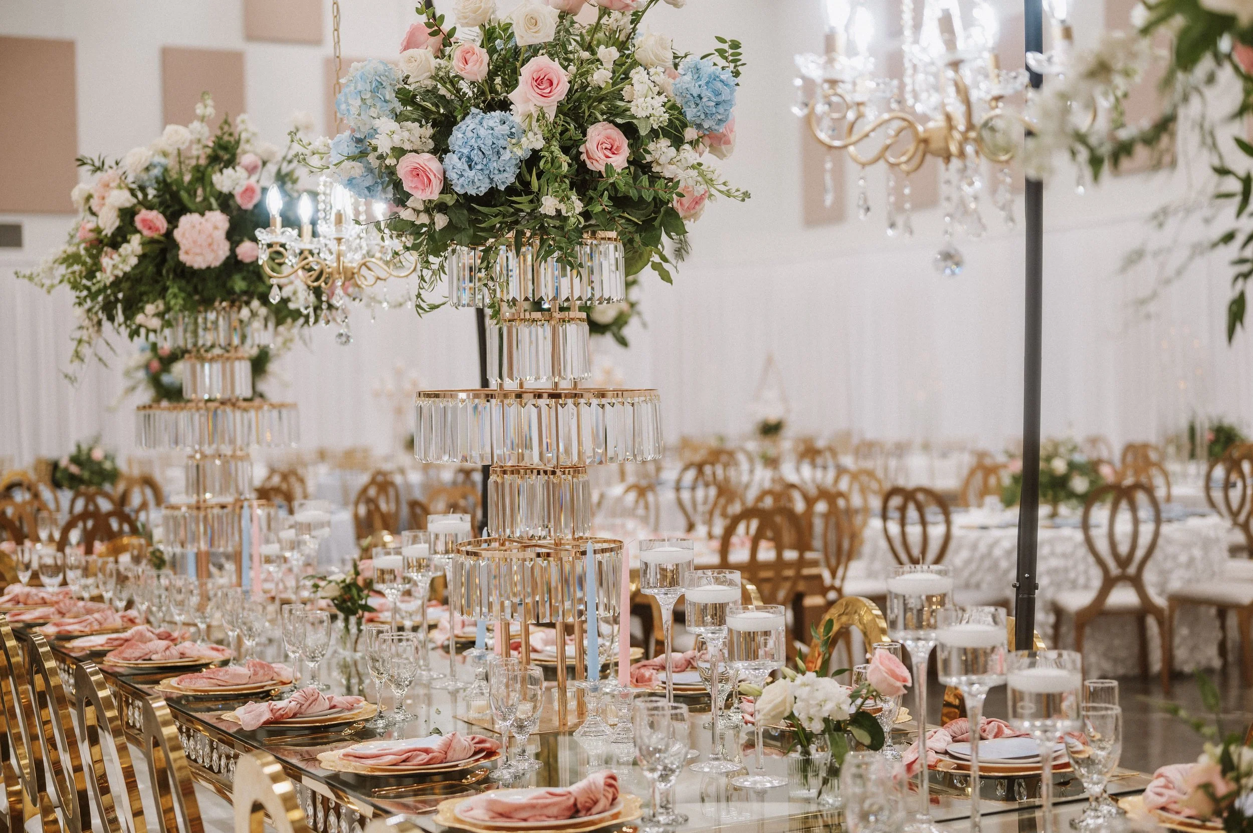 Elegant banquet table decorated with tall floral centerpieces featuring pink, white, and blue flowers, crystal chandeliers, and gold accents, set in a ballroom with rows of chairs.