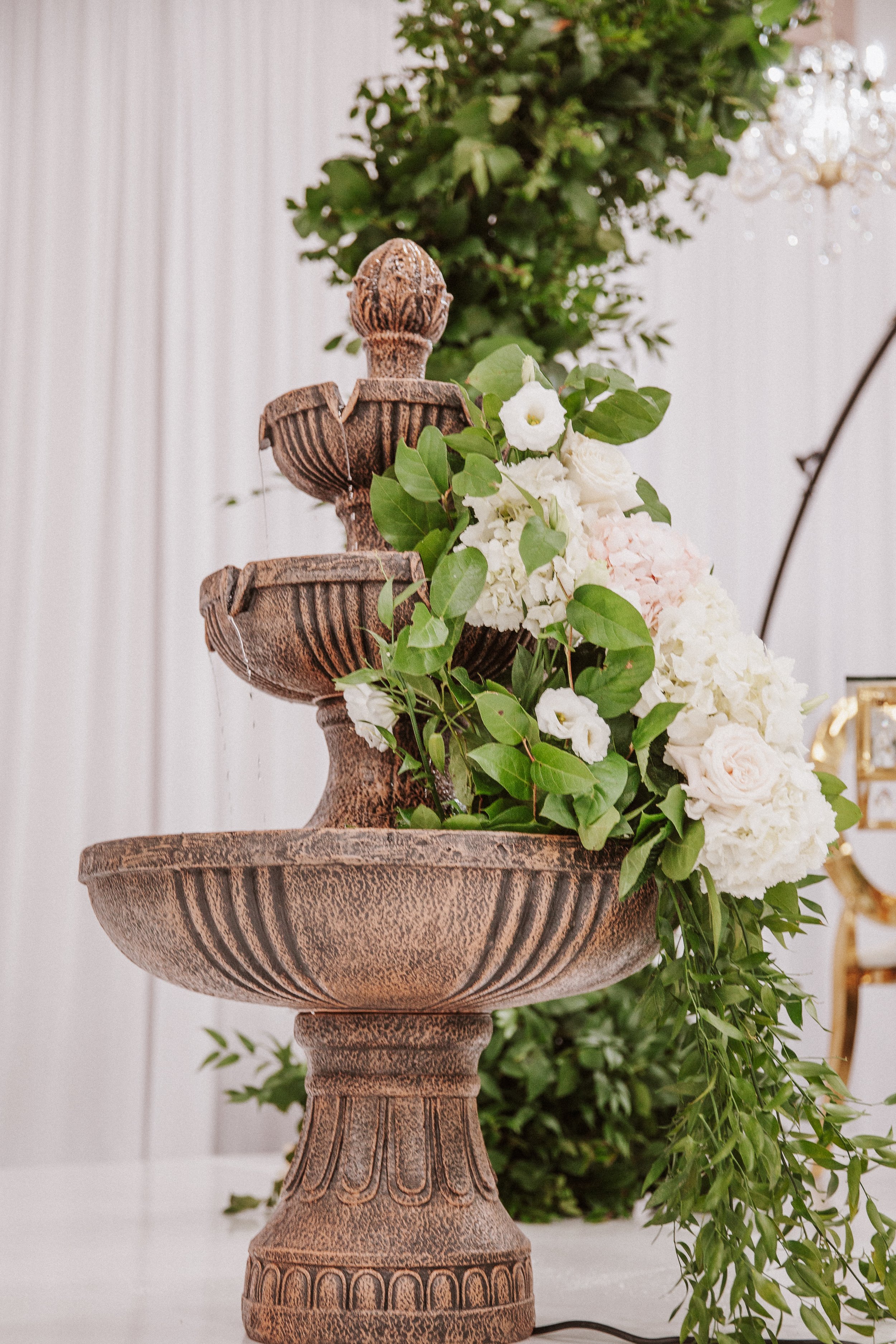 A decorative stone fountain with a bust sculpture on top, surrounded by white and pale pink flowers and green foliage, in an indoor setting with a white wall and a chandelier in the background.