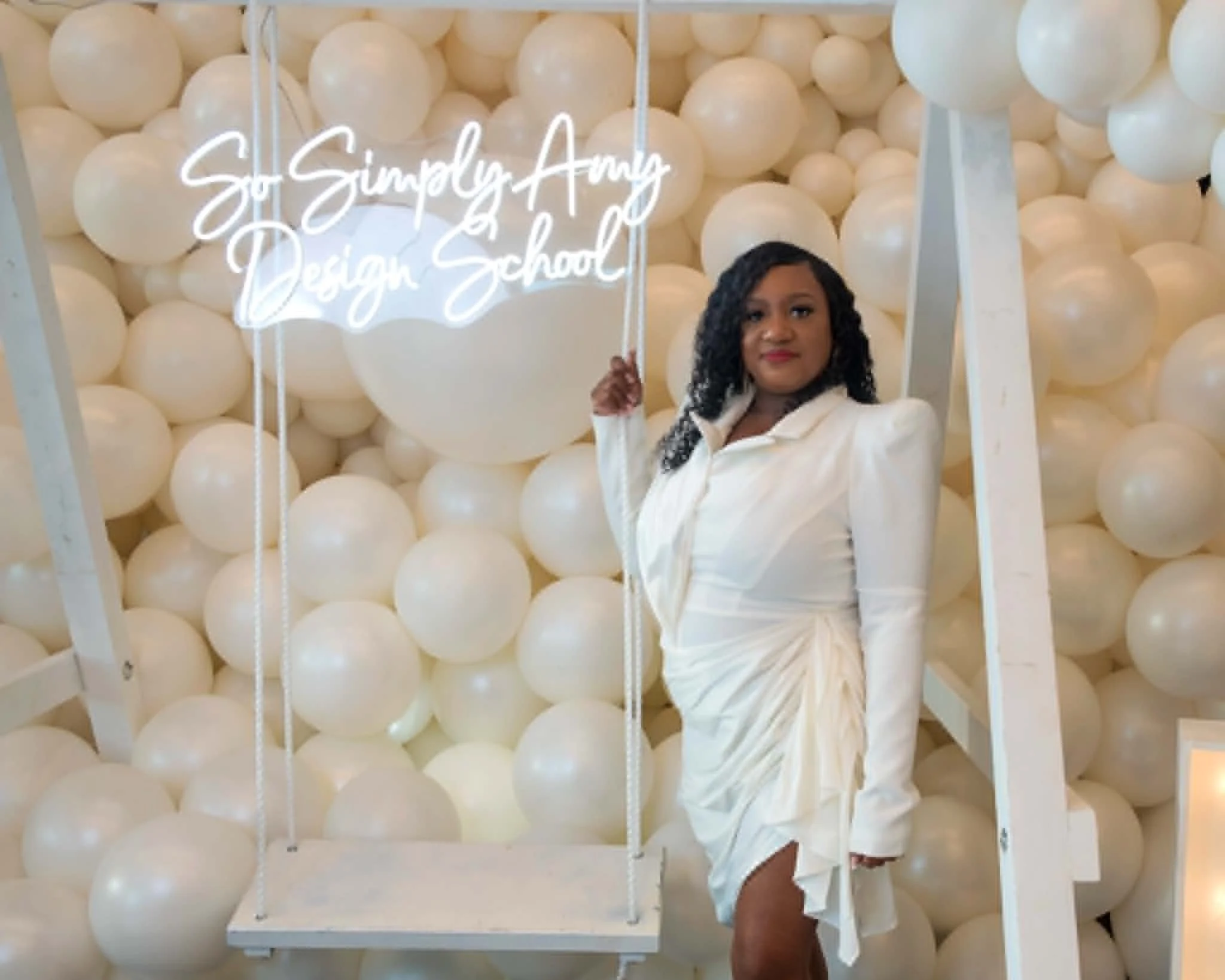 Woman in white blazer and skirt standing in front of a wall filled with white balloons, holding a neon sign that reads 'So Simply Amazing Design School'.