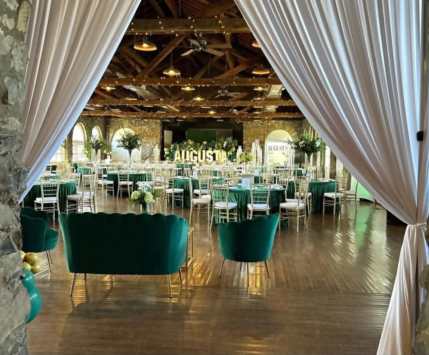 Elegant wedding reception hall decorated with green and white floral arrangements, tables with green tablecloths, and gold chairs. The hall features exposed brick walls, arched windows, and a decorated ceiling with string lights. The decor includes a sign that reads 'AUGUST'.