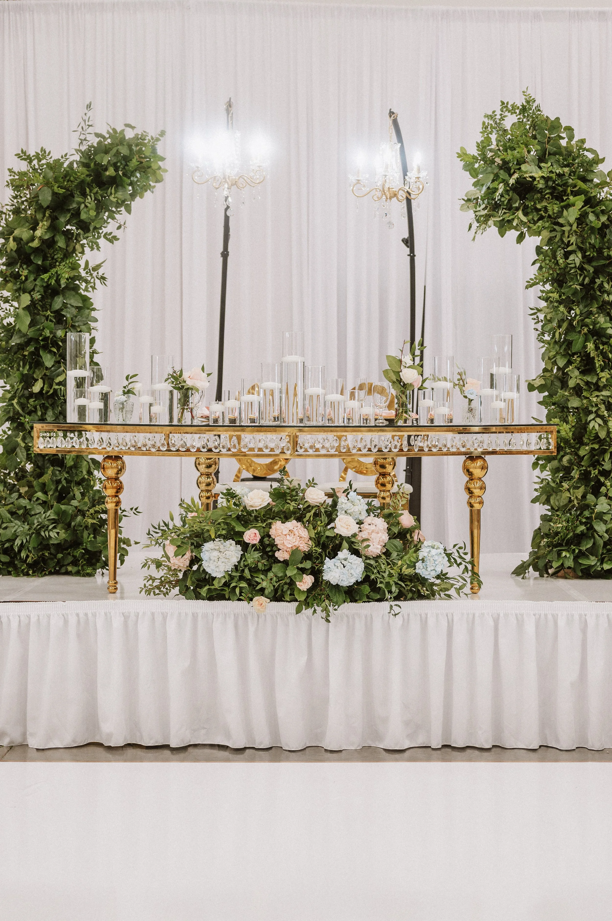 A decorated table with glass candle holders, small floral arrangements, and greenery, set against a white curtain backdrop, with large leafy green plants on either side.