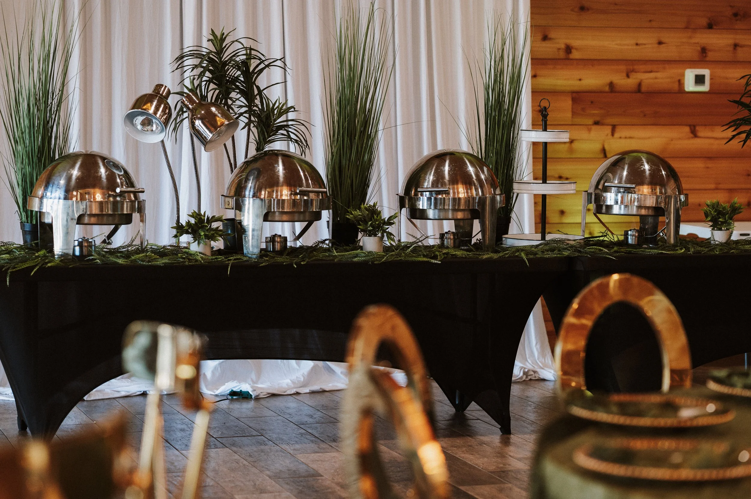 Buffet table setup with four chafing dishes, tall decorative grass and plants, tiered serving tray, and small potted plants on a black tablecloth in a wooden-walled room.
