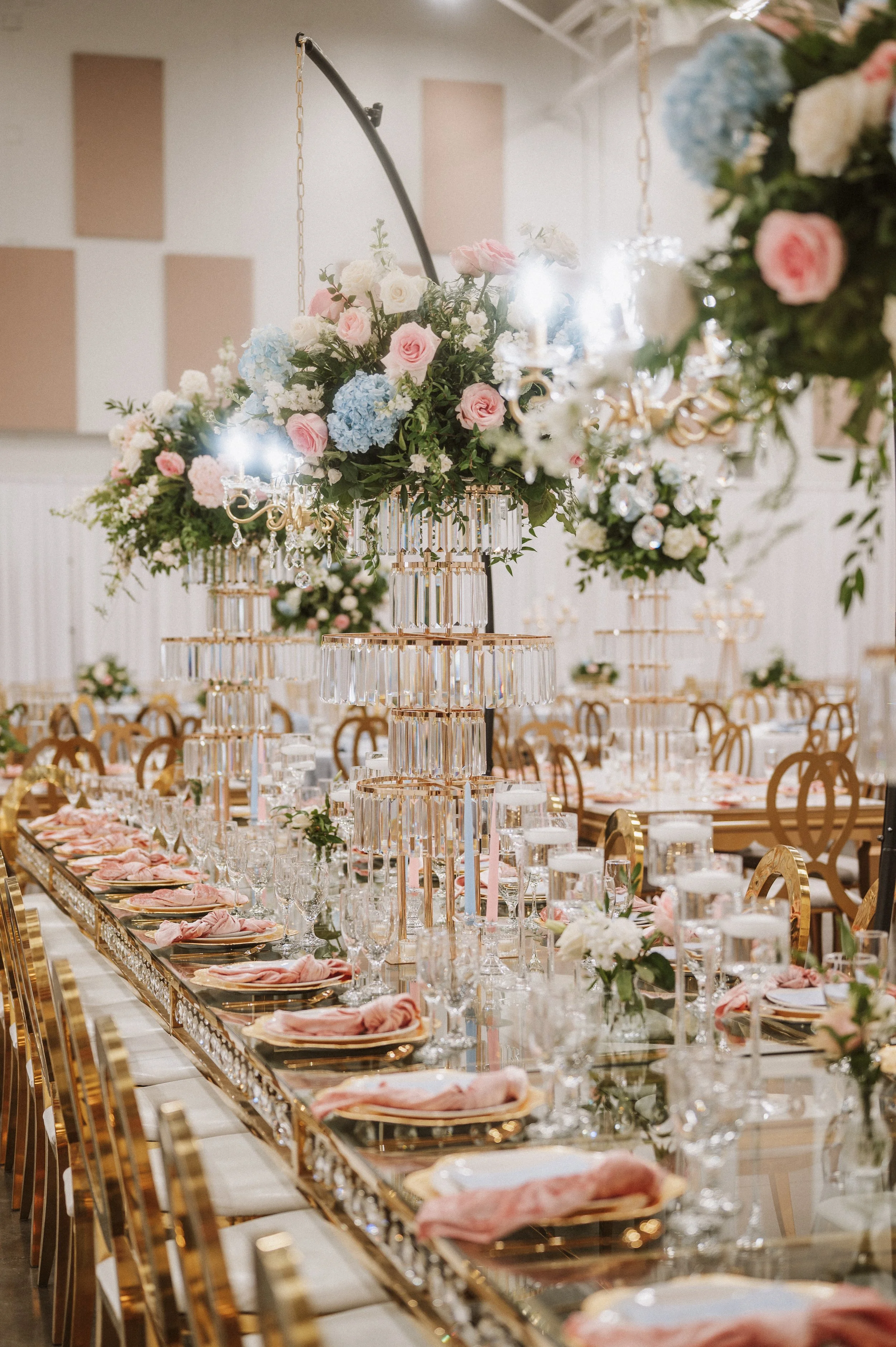 A luxurious banquet table decorated with pink, white, and blue flowers, with elegant gold chairs, crystal glassware, and pink napkins. Tall floral centerpieces with chandeliers hanging above.
