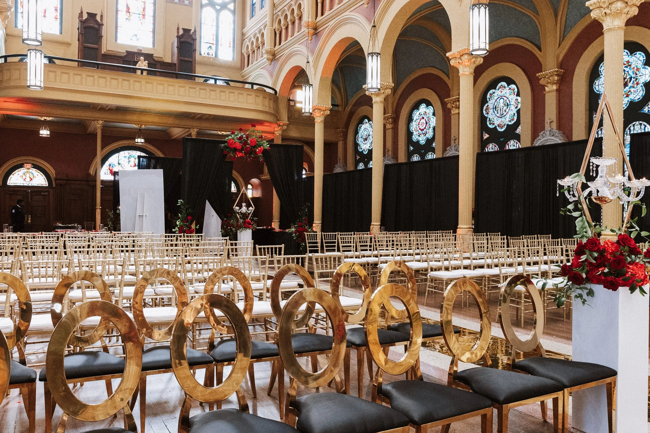Interior of a church or cathedral decorated for a wedding, with rows of gold chairs with black cushions, floral arrangements, and stained glass windows.