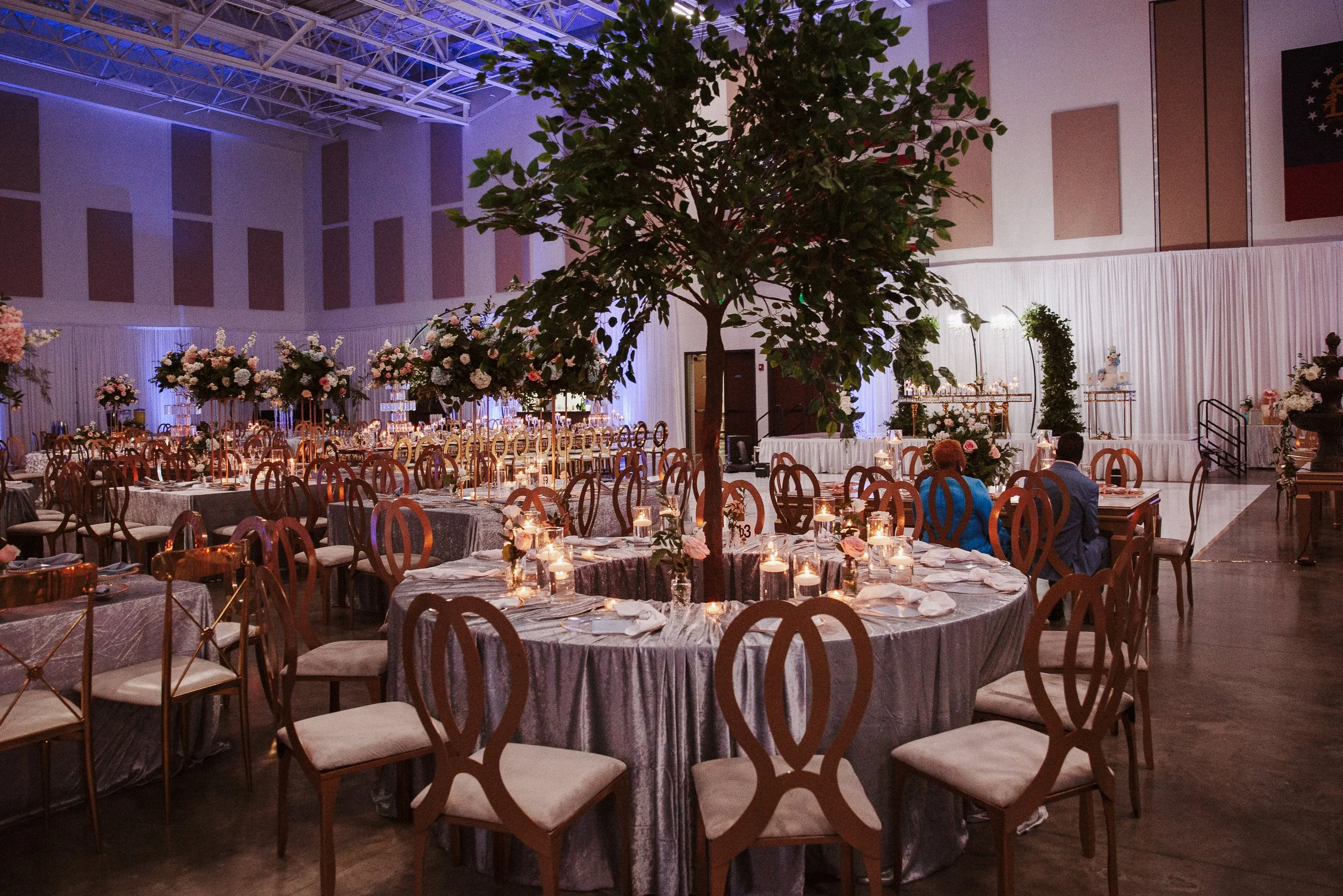 Inside a banquet hall decorated for an event with round tables covered with gray tablecloths, each set with plates, glasses, and lit candles. There is a large green tree in the center of the space and floral arrangements on the tables. The hall has high ceilings with exposed beams and is illuminated with purple and blue lighting.