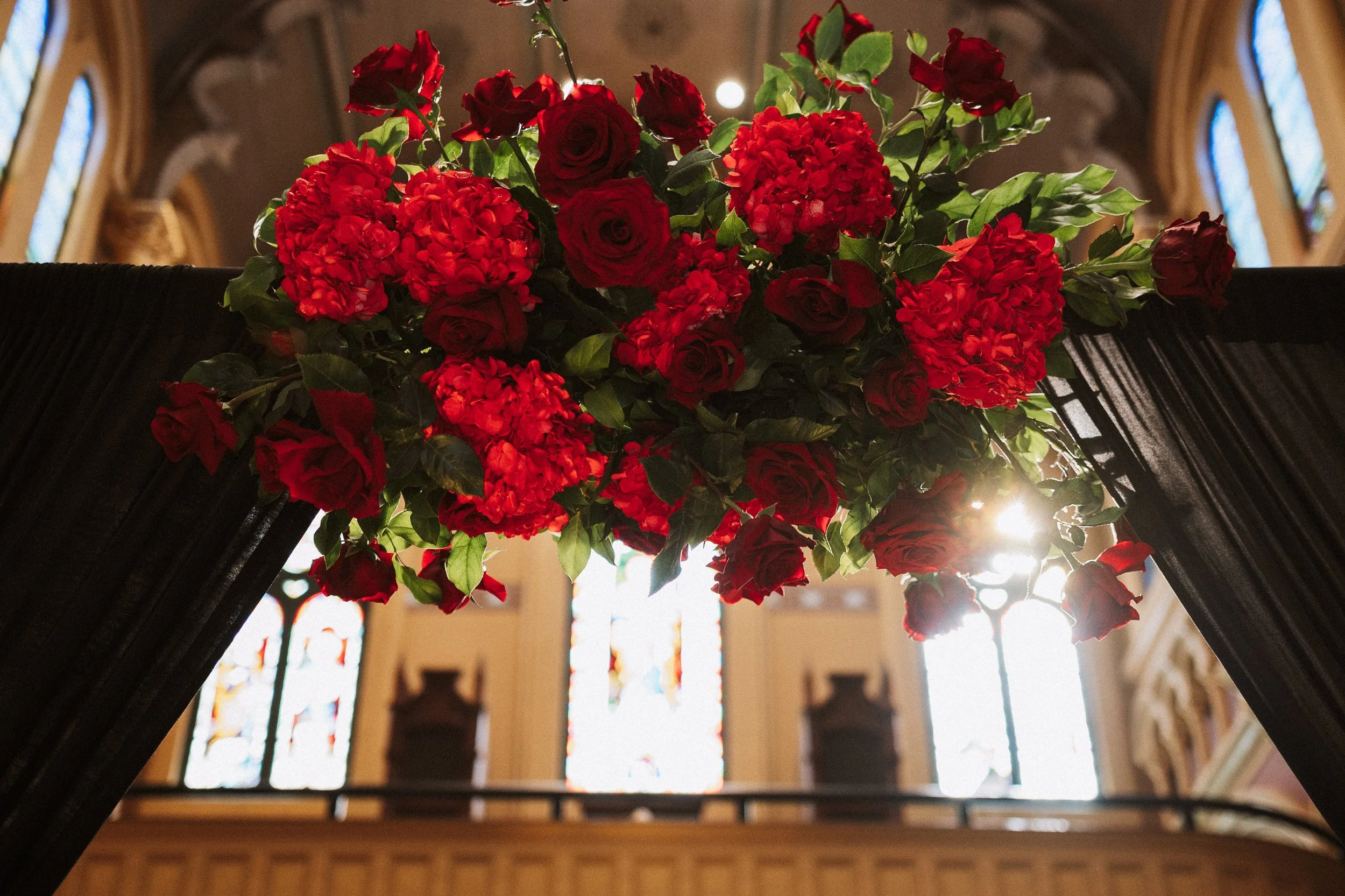 A vibrant floral arrangement featuring red roses, hydrangeas, and greenery, placed on a black surface, with stained glass windows in the background and sunlight streaming through.