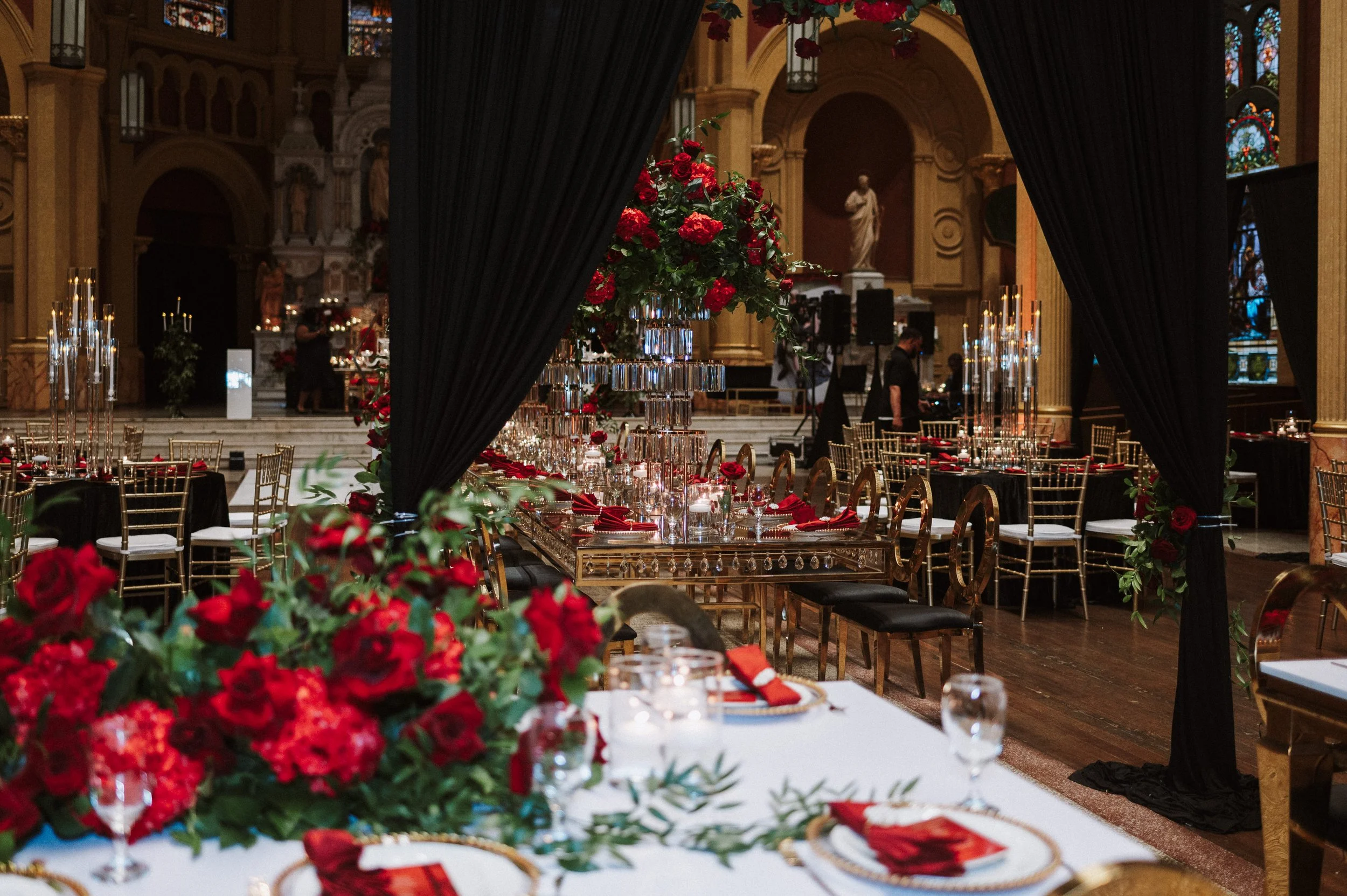 Elegant event setup inside a church with long tables decorated with red flowers, gold chairs, and candles, partially obscured by black curtains, with stained glass windows and religious statues in the background.