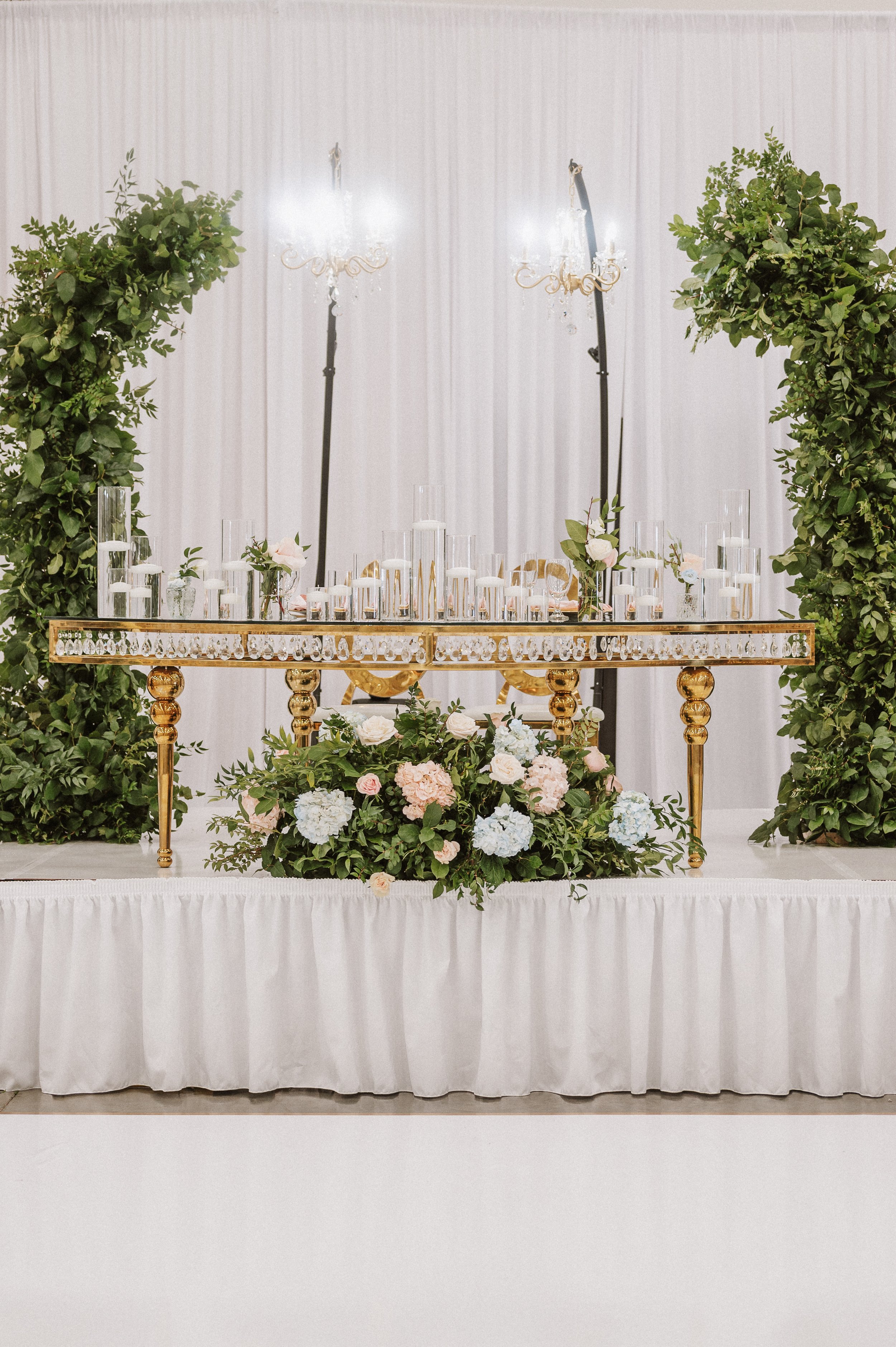 Elegant reception table decorated with white and pink flowers, greenery, glass candle holders, and gold accents, set against a white backdrop with greenery on either side.