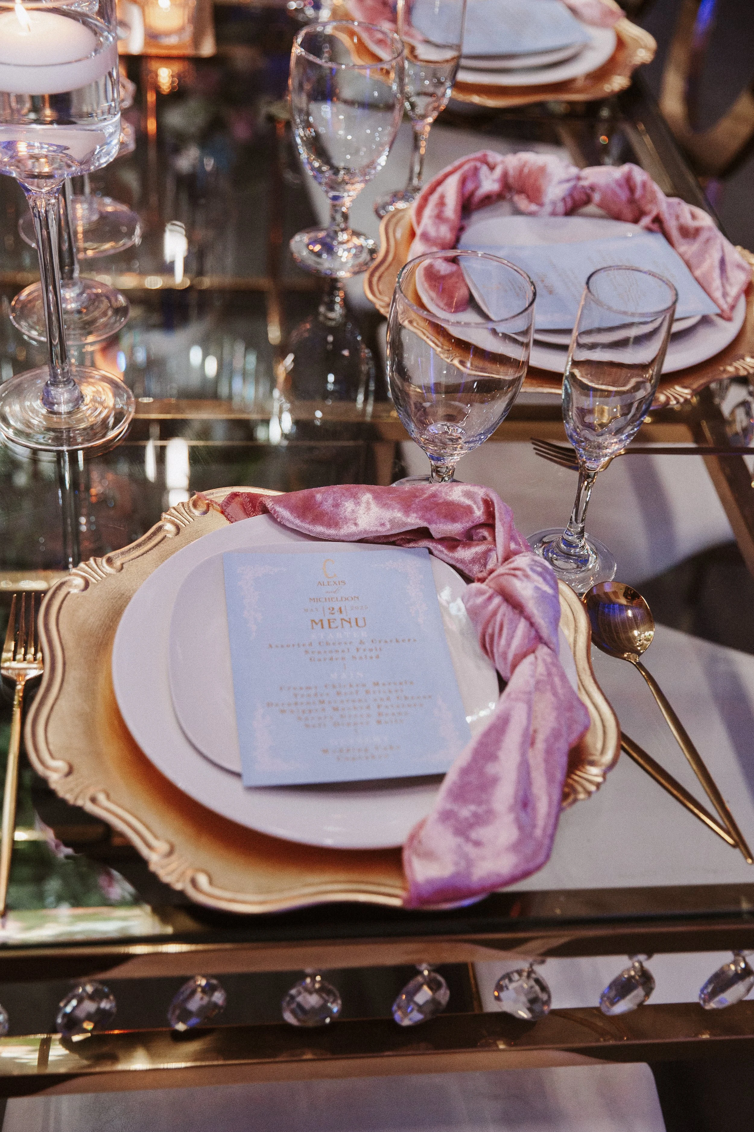 Elegant table setting with gold-rimmed plates, pink velvet napkins, multiple wine glasses, water glasses, and a menu card, all arranged on a glass-topped table with crystal embellishments.