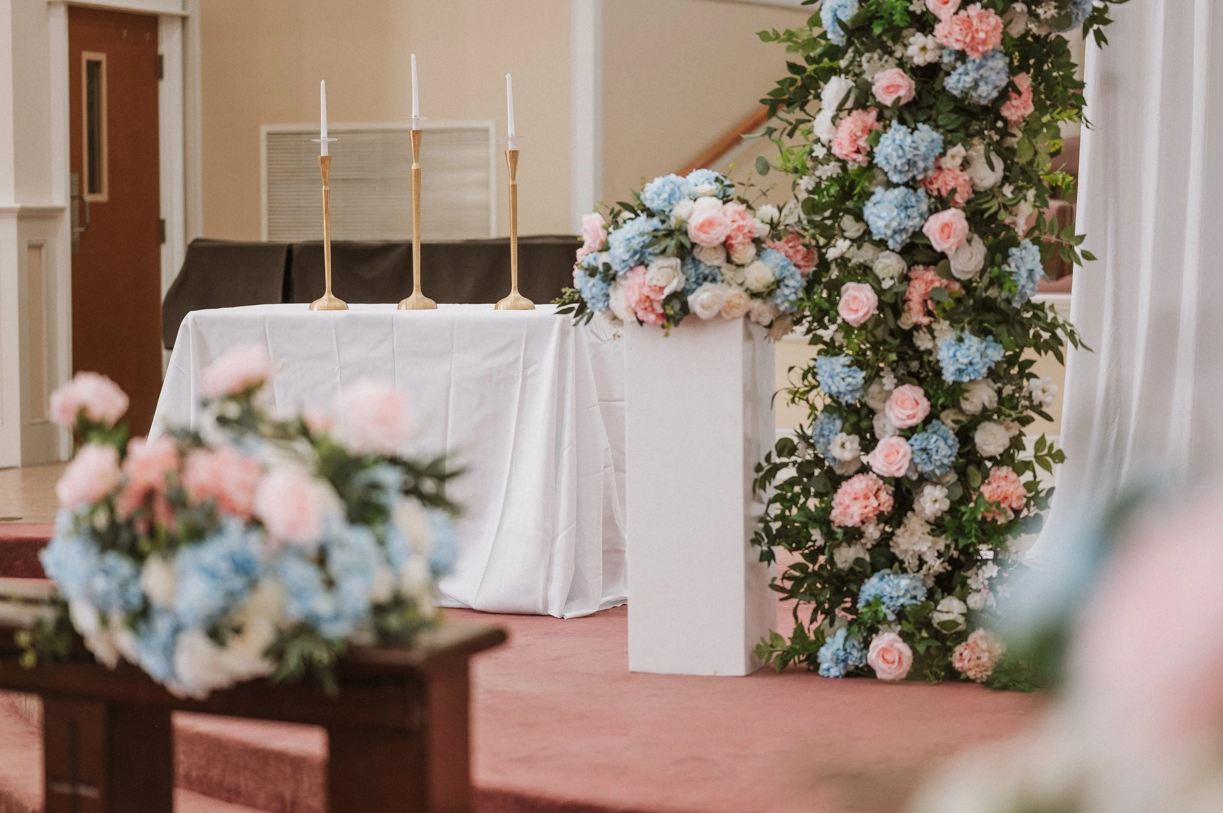 A wedding or event setup with a table covered in a white cloth, decorated with tall candle holders and a large floral arrangement featuring pastel-colored roses and hydrangeas, with additional flowers and greenery.