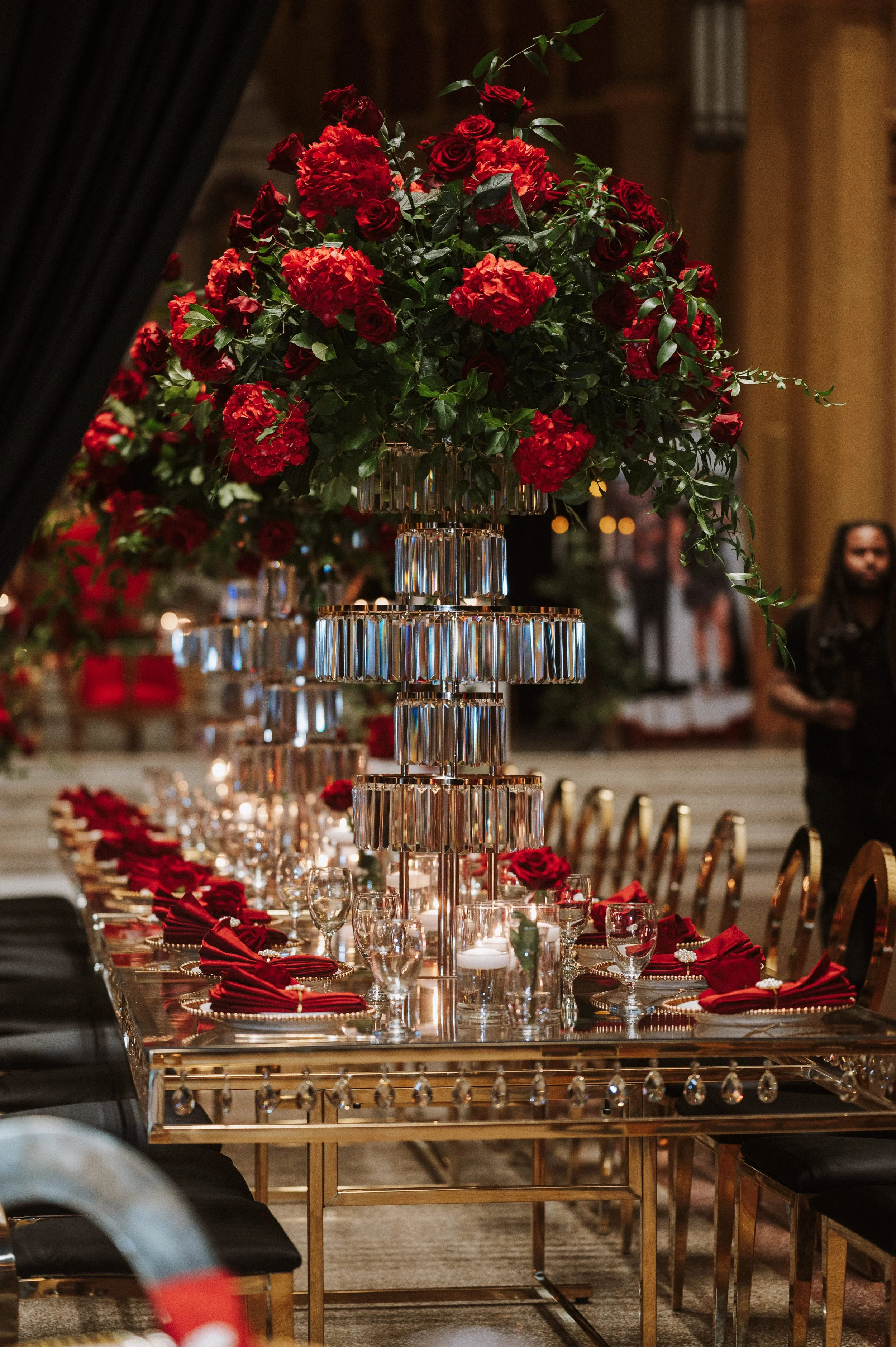 Elegant banquet table set with red napkins, crystal glassware, and tall floral centerpiece of red roses and hydrangeas with green foliage, in a dimly lit, sophisticated dining room.