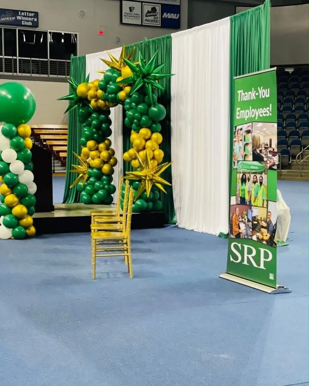Decorative stage with an arch made of green, yellow, and white balloons, a 'Thank-You Employees!' sign, and a yellow chair in front.
