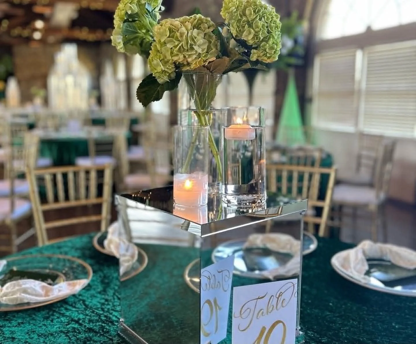 A table centerpiece with green hydrangea flowers in a clear glass vase, two floating candles in water-filled glass holders, placed on a reflective metallic platform, in a decorated restaurant with chairs, plates, and napkins in the background.