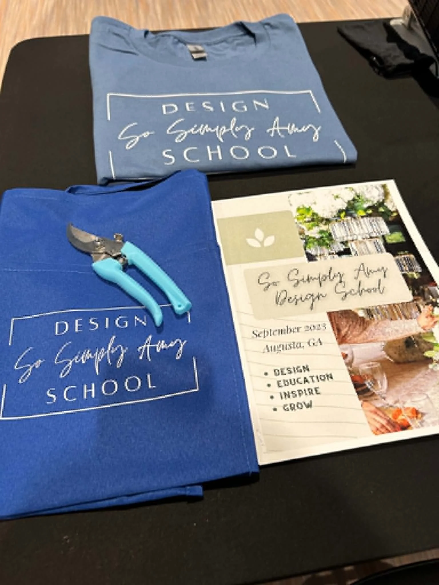 A blue t-shirt with white text reading "Design, So Simply Amazing, School" and a pamphlet with the same text, along with a pair of small multicolored pliers on a black table.