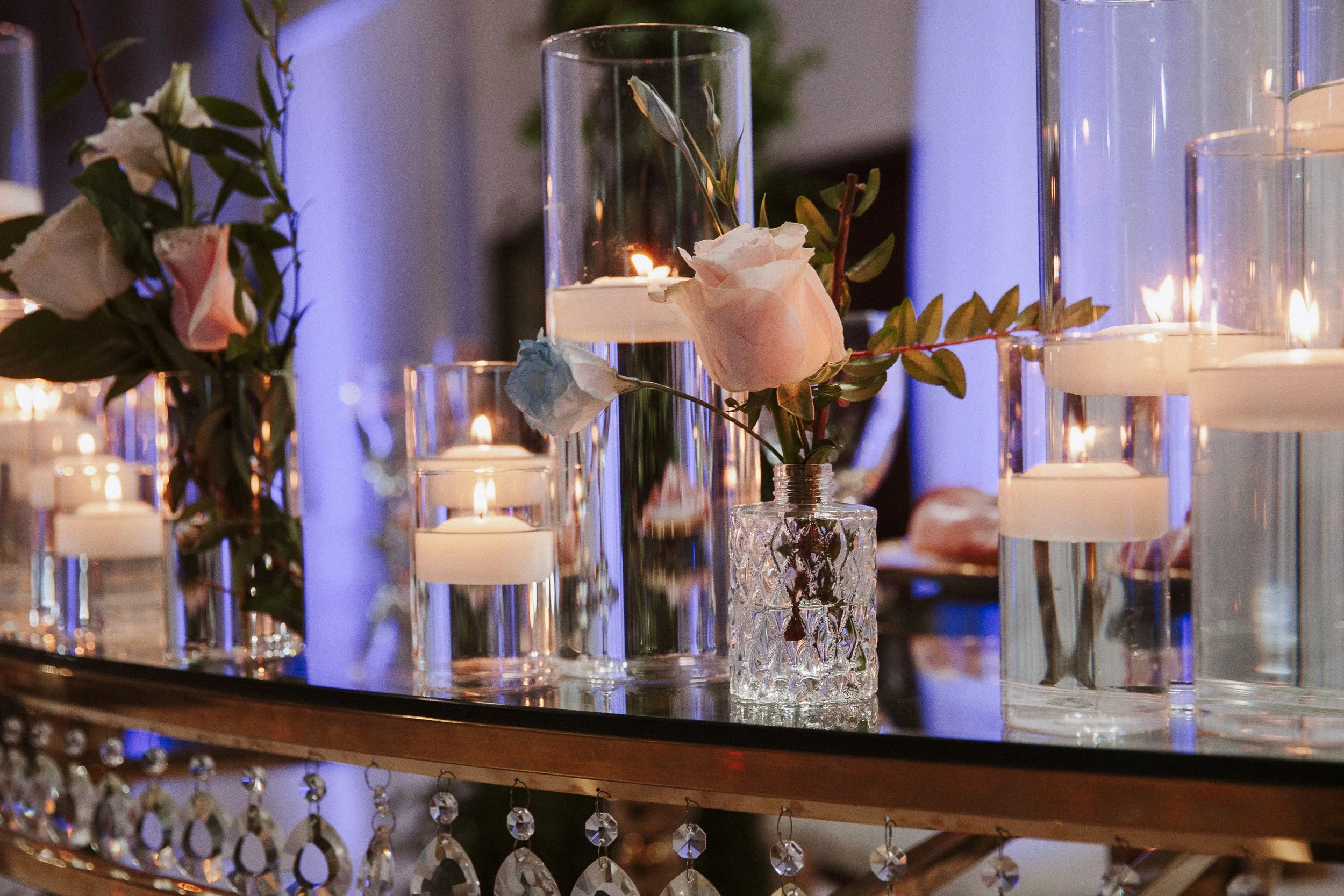 Decorative table centerpiece with candles in glass holders and pink and white roses with green foliage, illuminated by soft lighting.