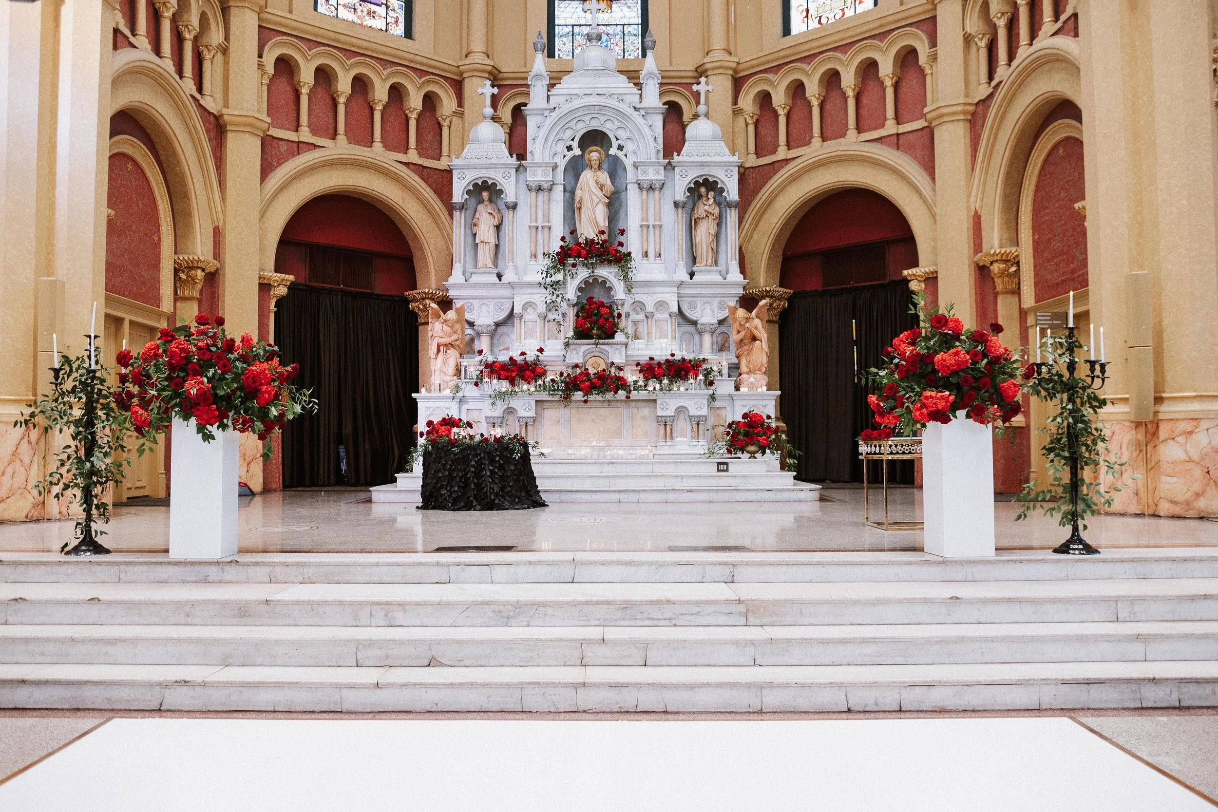 Inside a church, an altar decorated with red flowers and religious statues, with stained glass windows in the background and marble steps leading up to it.