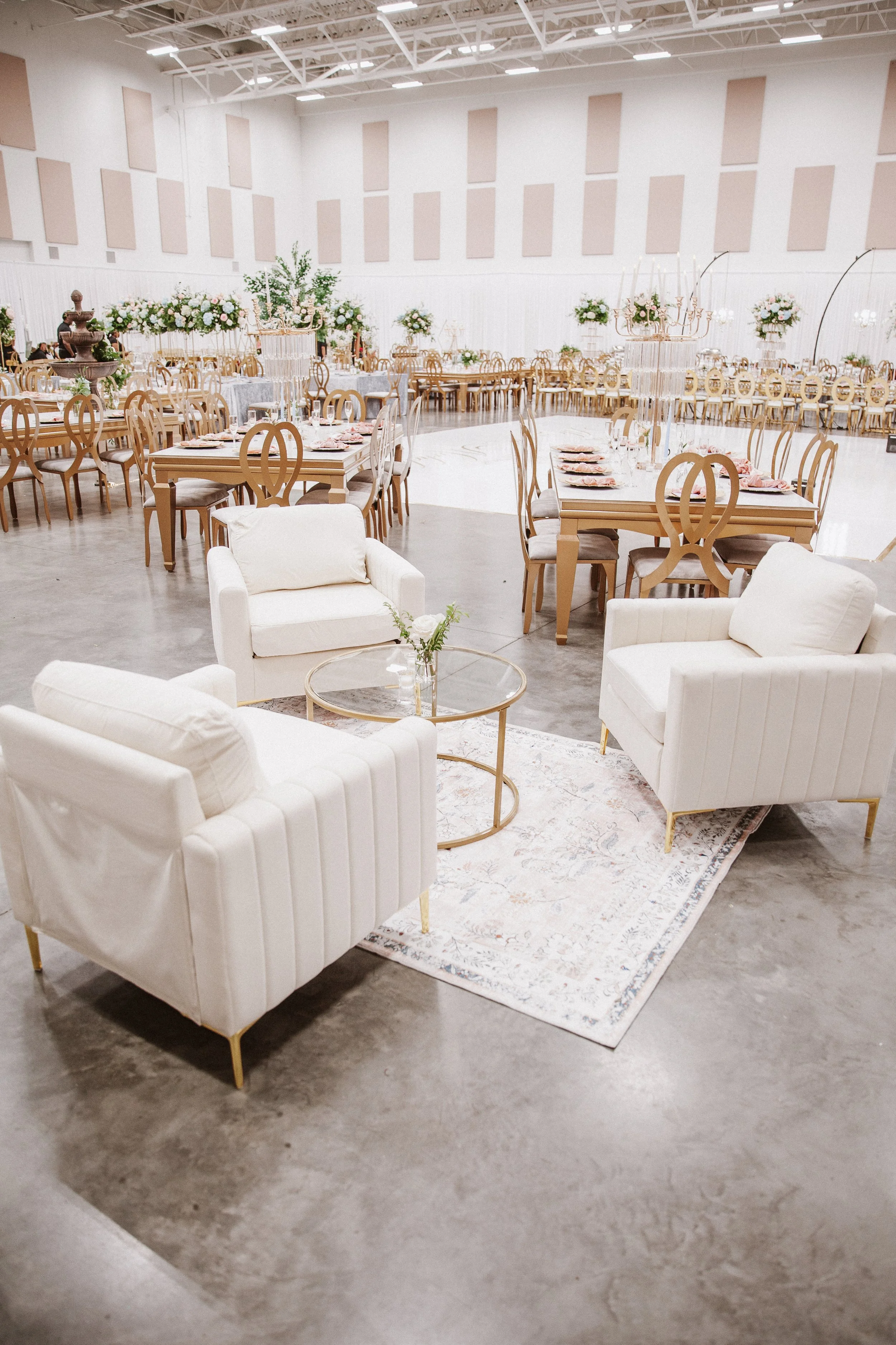 Empty banquet hall decorated with light-colored wooden chairs arranged around long tables with pink napkins and floral centerpieces. White sofas are in the foreground, and the hall has high ceilings with exposed beams.