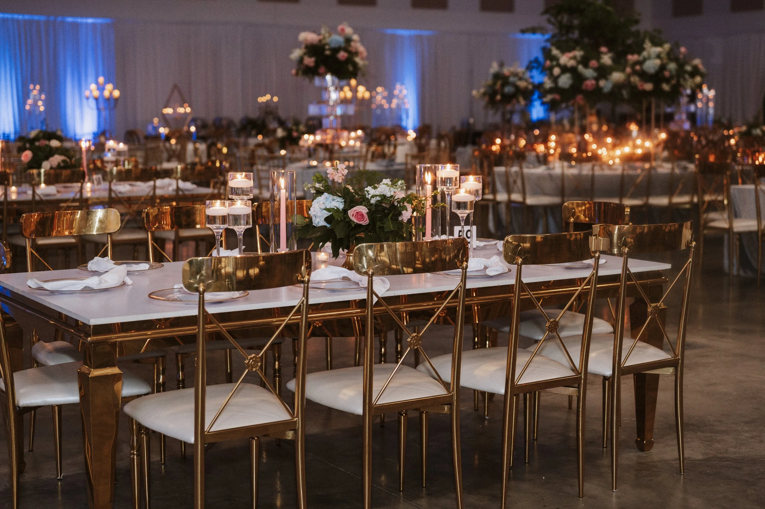 Elegant event hall decorated for a celebration with tall floral centerpieces, candles, and gold chairs arranged around tables.