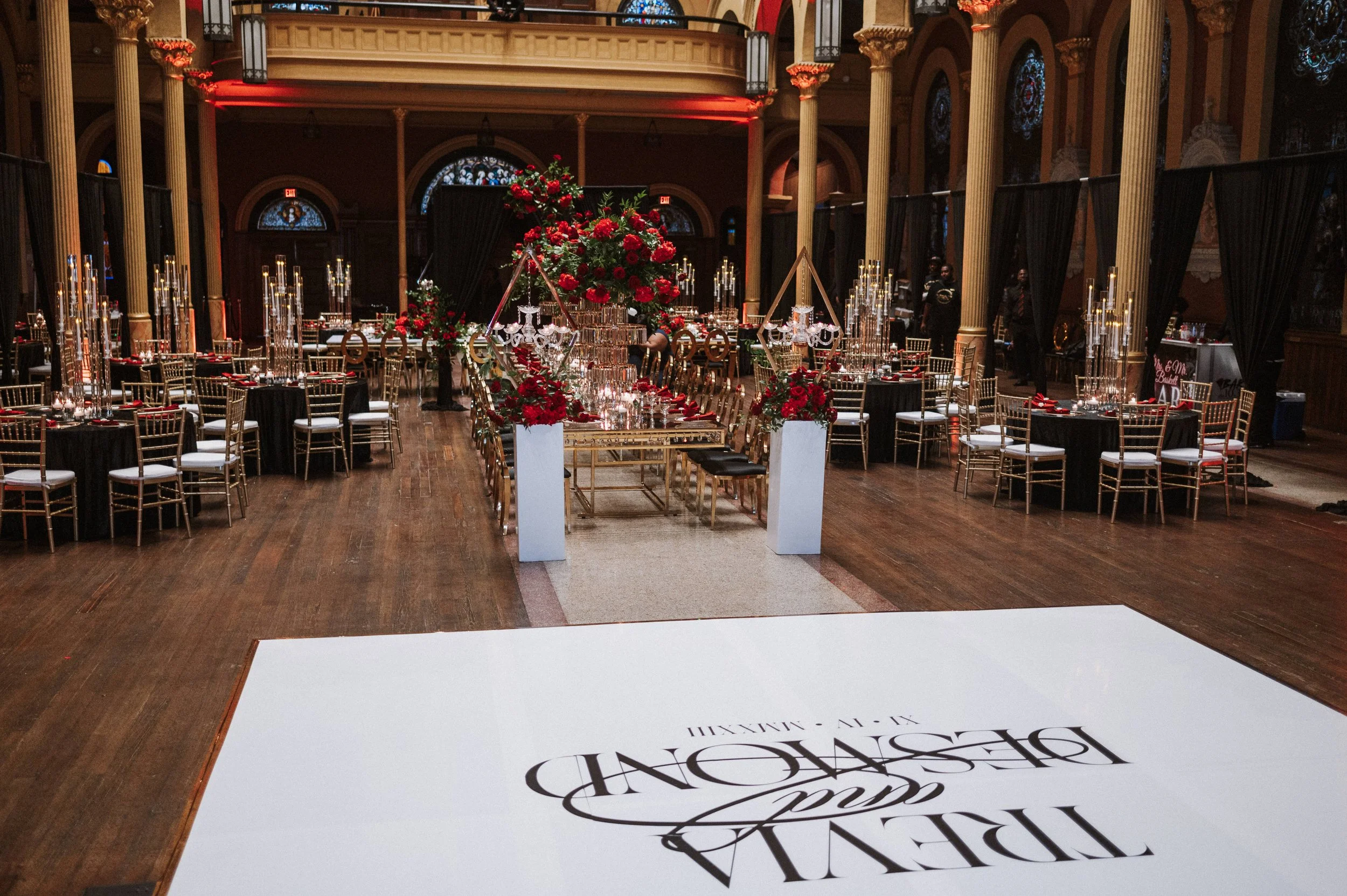 A decorated banquet hall with tables, chairs, and floral arrangements, set up for an event in a grand, ornate room with stained glass windows and high ceilings.