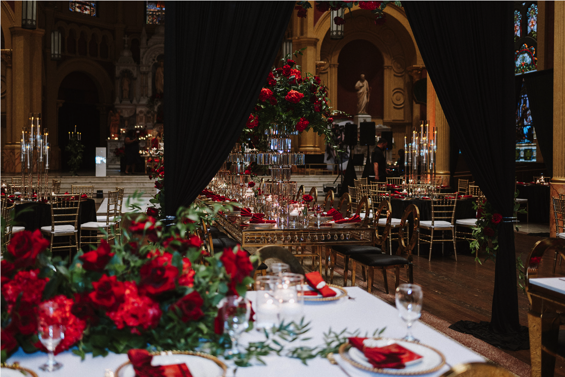 Elegant banquet hall decorated with black curtains, red floral arrangements, gold chairs, and candle centerpieces for a formal event.
