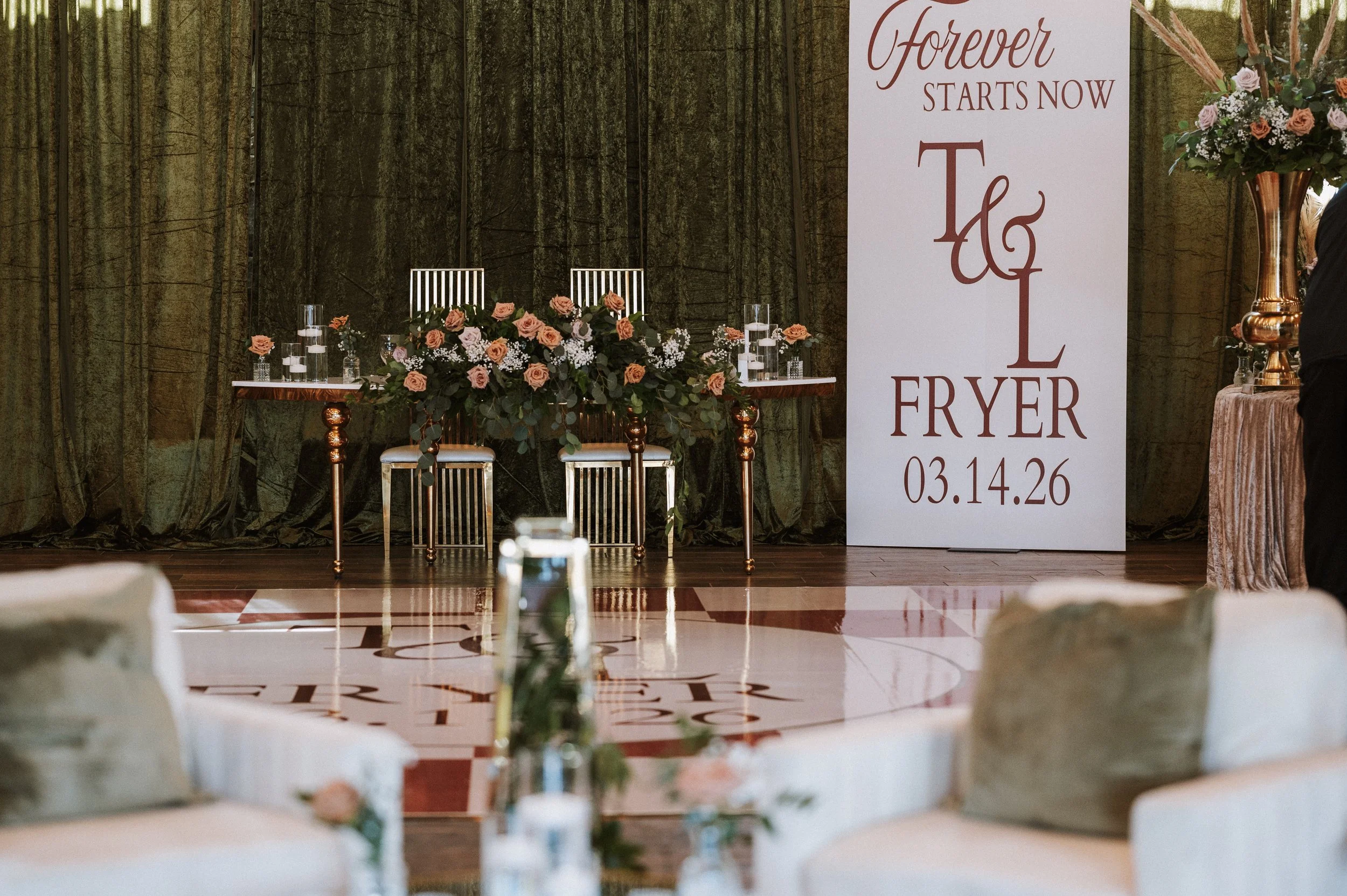 Wedding reception setup with a large white sign displaying "Forever Starts Now" and the names and date. Floral arrangements on tables at the front and behind the sign. Chairs and tables for guests. Draped curtains and dark green backdrop.