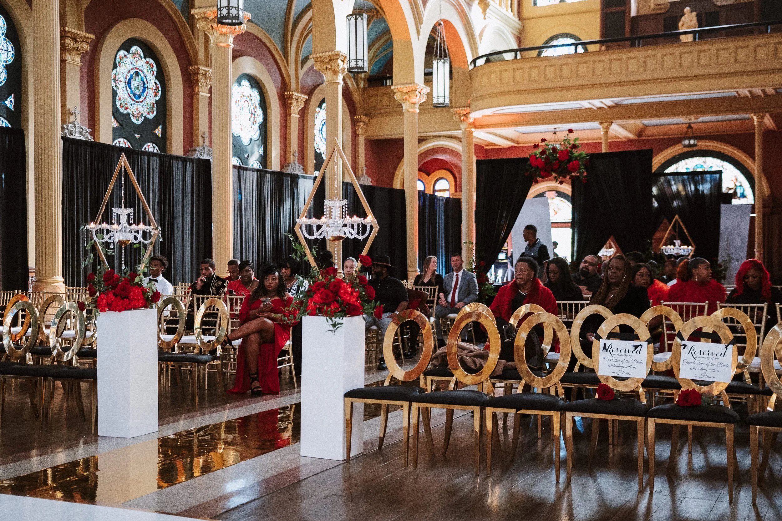 Interior of a church decorated for a wedding, with seated guests, floral arrangements, chandeliers, stained glass windows, and reserved signs on some seats.