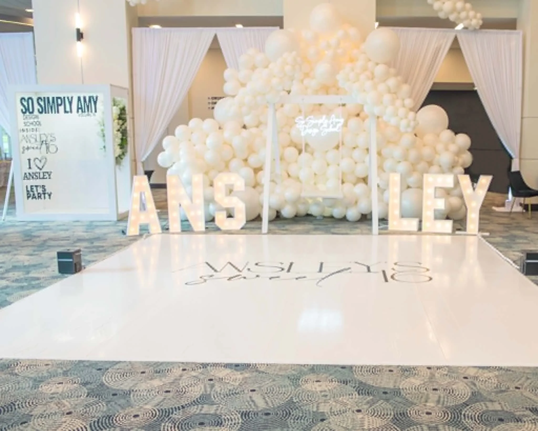 A decorated event space with a large white ball and fabric backdrop, illuminated letter lights spelling 'LOVE,' and a white sign with blue text about a celebration of Ashley.