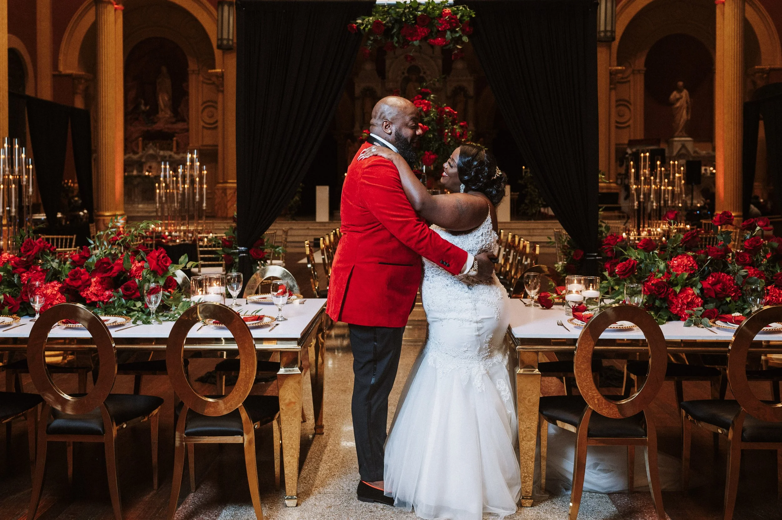 A wedding couple, a man in a red blazer and a woman in a white wedding dress, dancing and smiling in a decorated banquet hall with red roses and candle centerpieces.