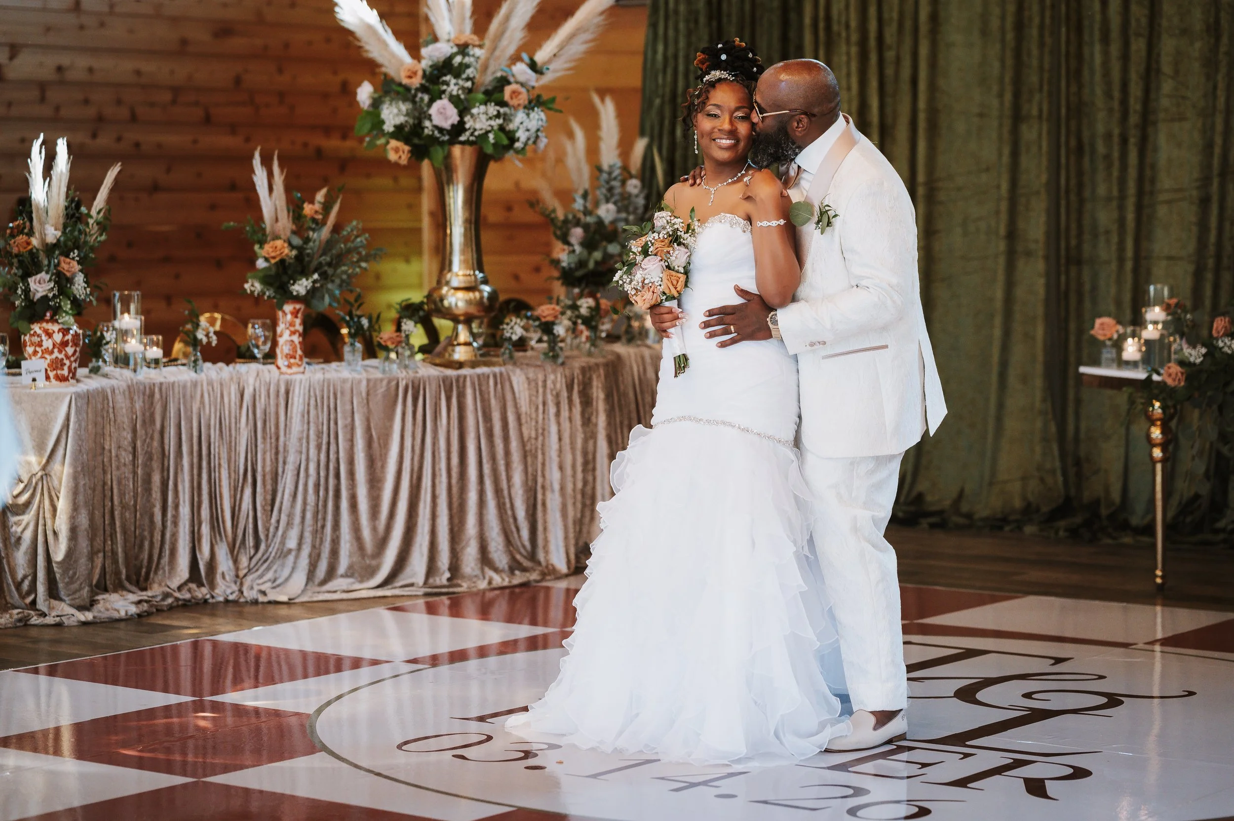 A bride and groom dancing at their wedding reception, surrounded by floral arrangements and a decorated table in a rustic indoor venue.