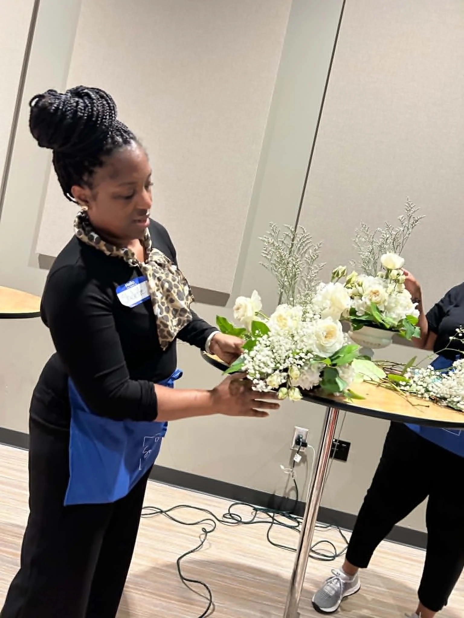 Woman wearing a black jacket and a leopard print scarf hands a bouquet of white flowers to another person during an indoor event.