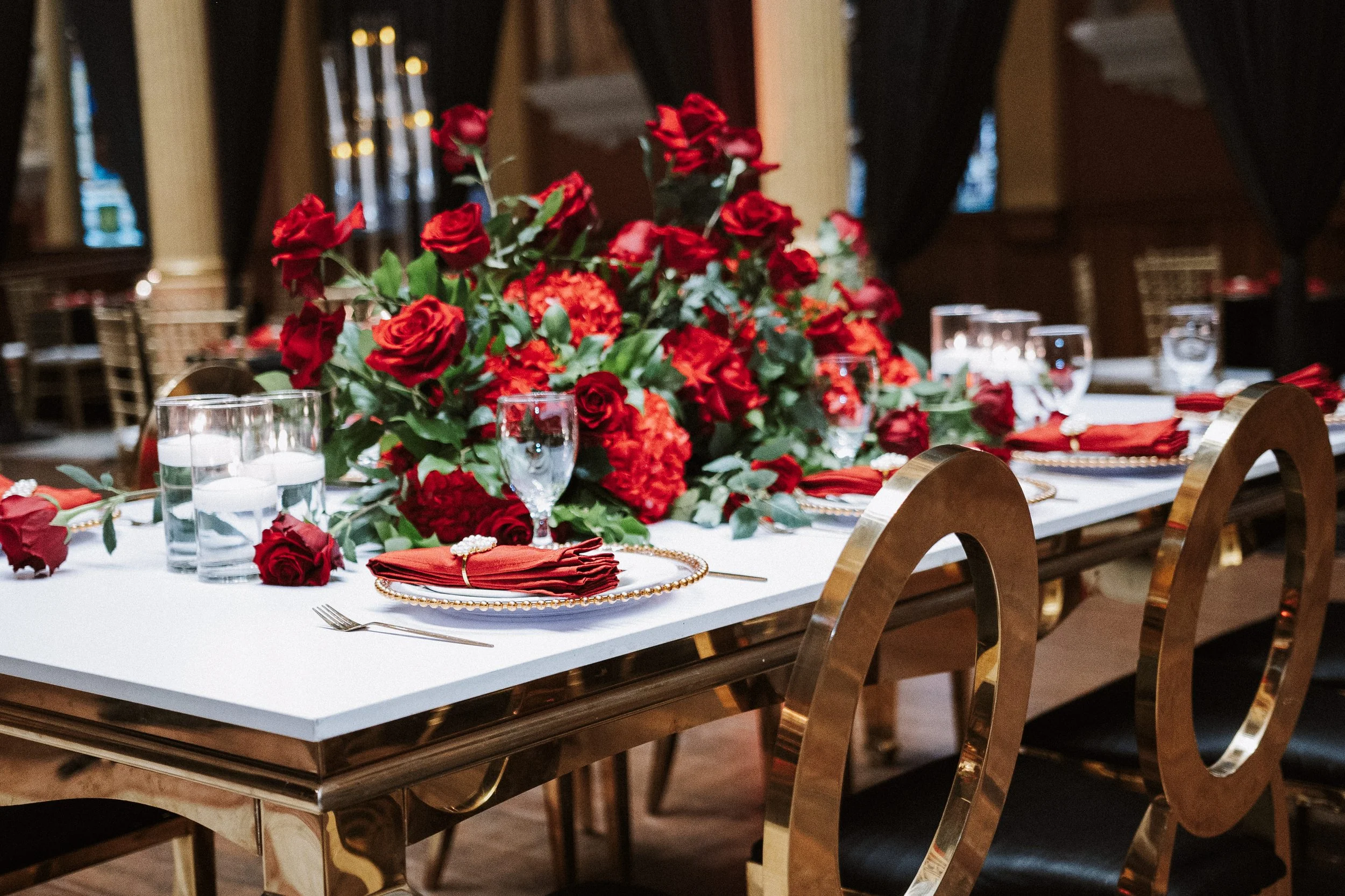A decorated banquet table with a large floral centerpiece made of red roses and other red flowers, set in a room with dark curtains and ornate wood and gold chairs, ready for an event or celebration.