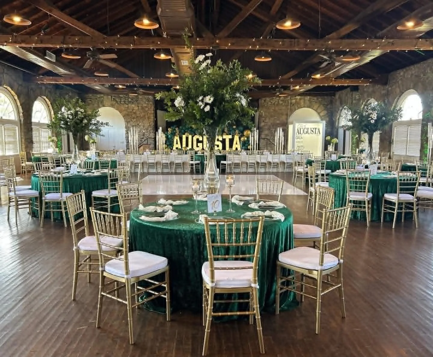 Indoor event space decorated for a celebration with green and gold themed tables, large floral arrangements, and golden chairs. There is a large floral centerpiece in the middle of the room and a sign with the word 'AUGUSTA' in the background.