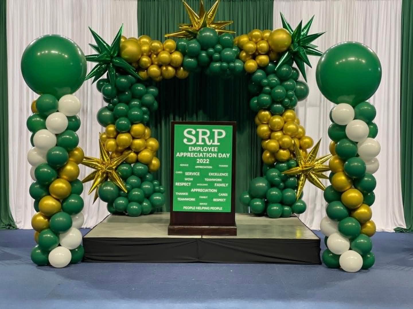 Balloon arch with green, gold, and white balloons at an employee appreciation event, with a sign in the center that reads 'SRP Employee Appreciation Day 2022'